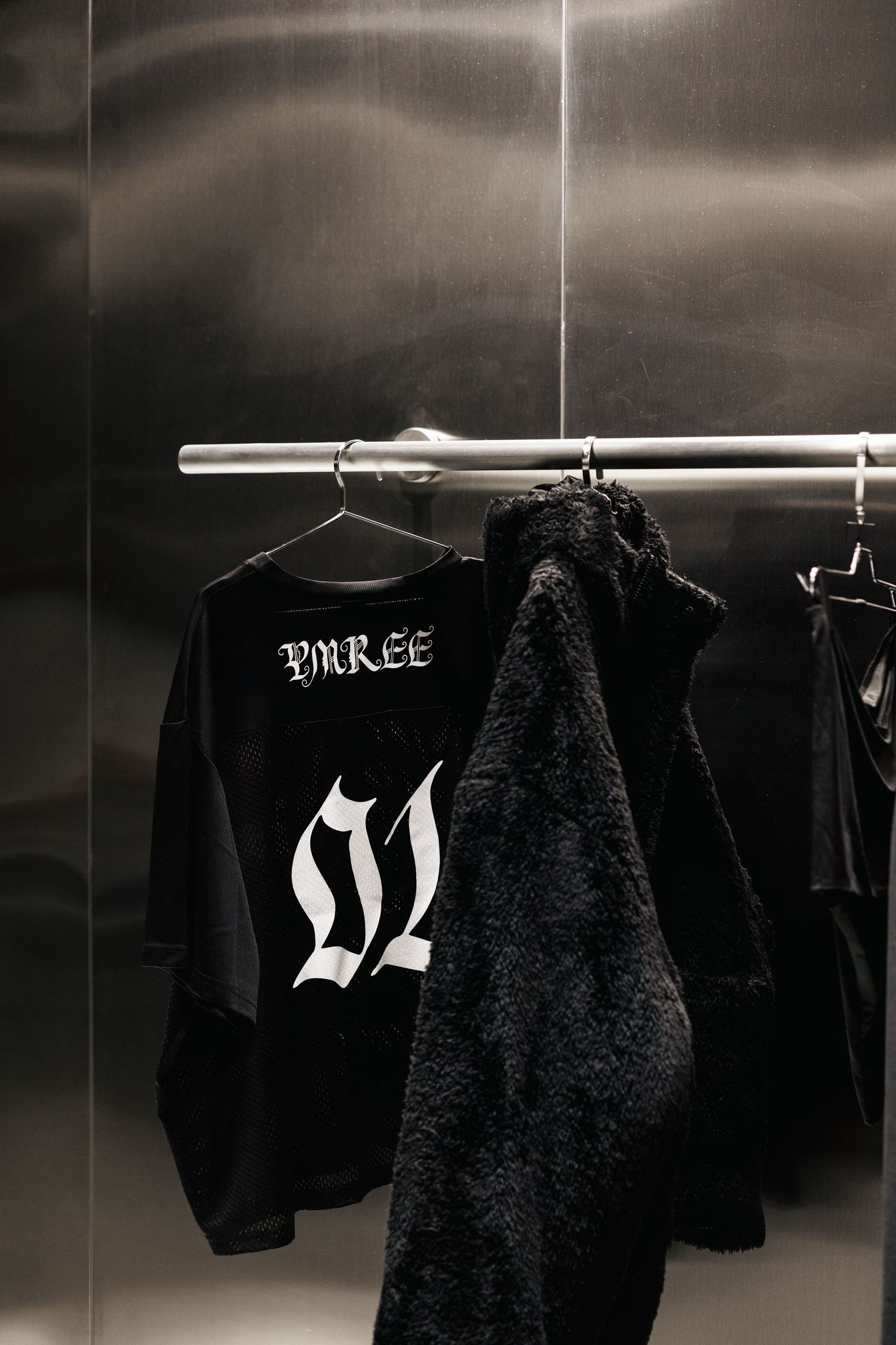 YMREE 01 gothic font black oversized tee and fur jacket displayed on hangers against metallic silver wall.