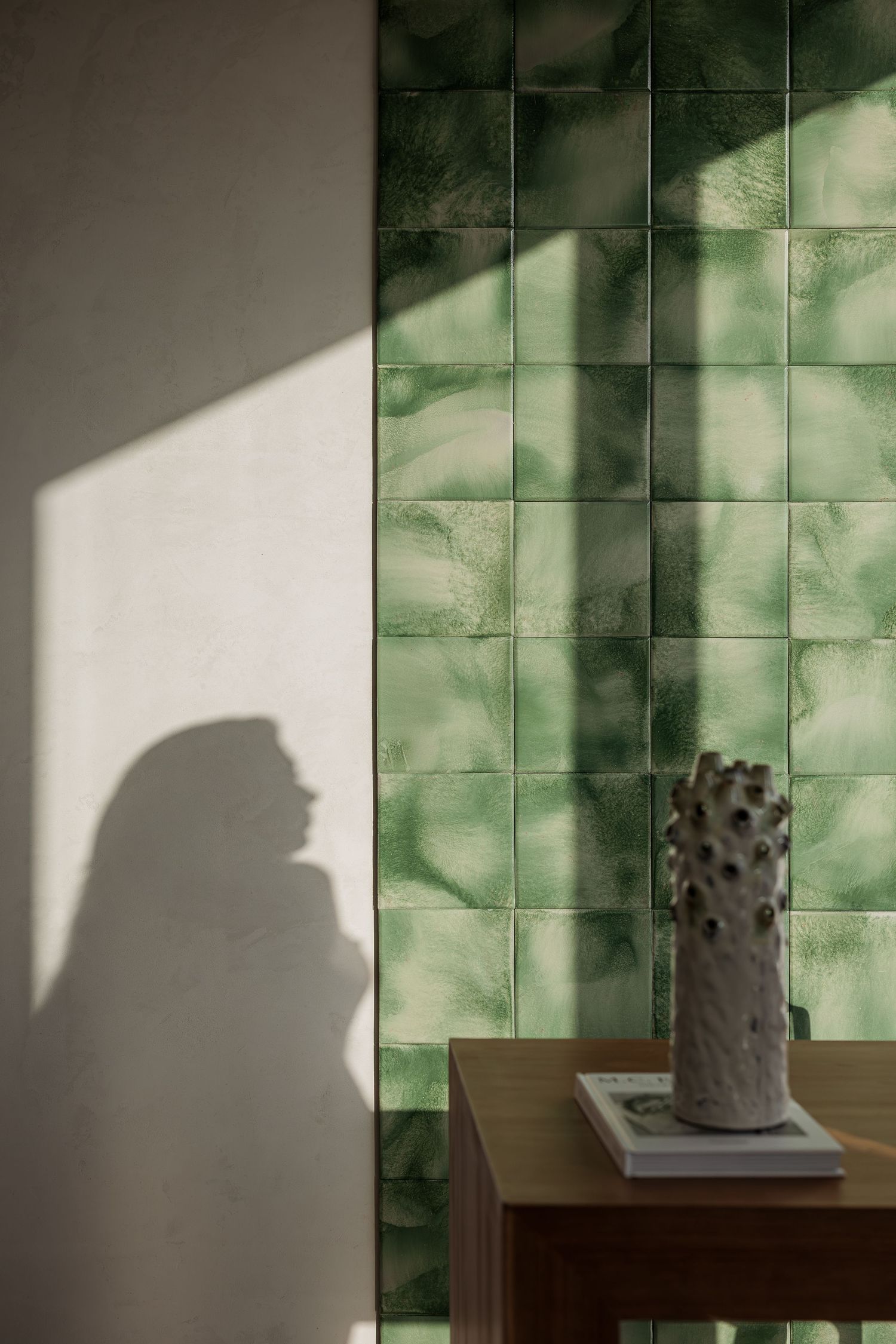 Green glazed ceramic tiles with shadow silhouette of woman and textured ceramic vase on wooden table.
