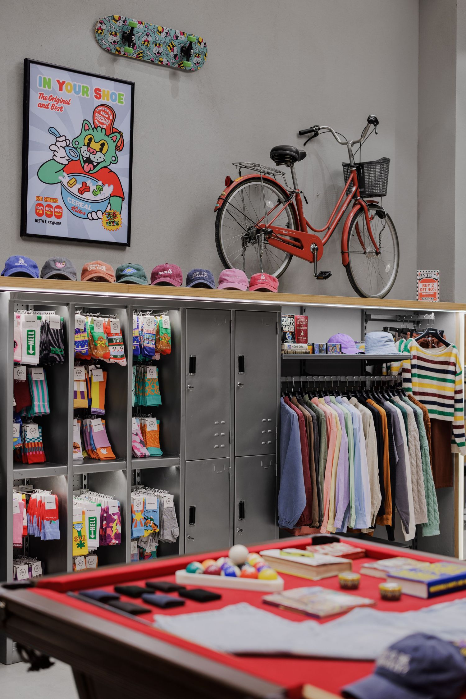 Trendy retail store interior featuring colorful socks, caps, striped shirts, lockers, a red bicycle, and a pool table.