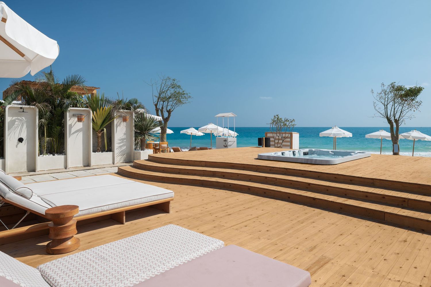 Upscale beachfront deck with white sunbeds, outdoor showers, umbrellas, and stunning ocean views.