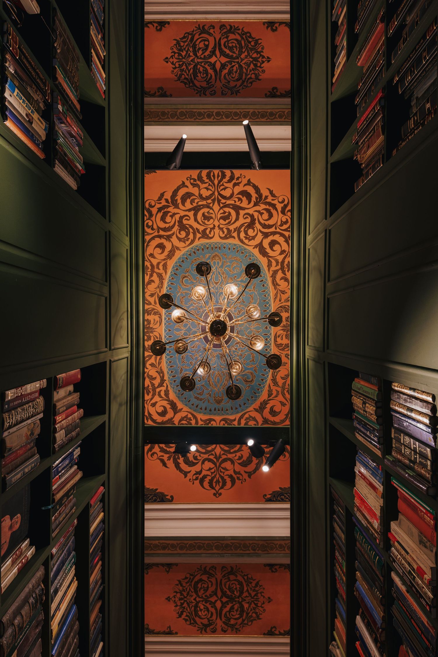 Looking up at ornate painted ceiling with chandelier surrounded by tall bookshelves in a grand library.