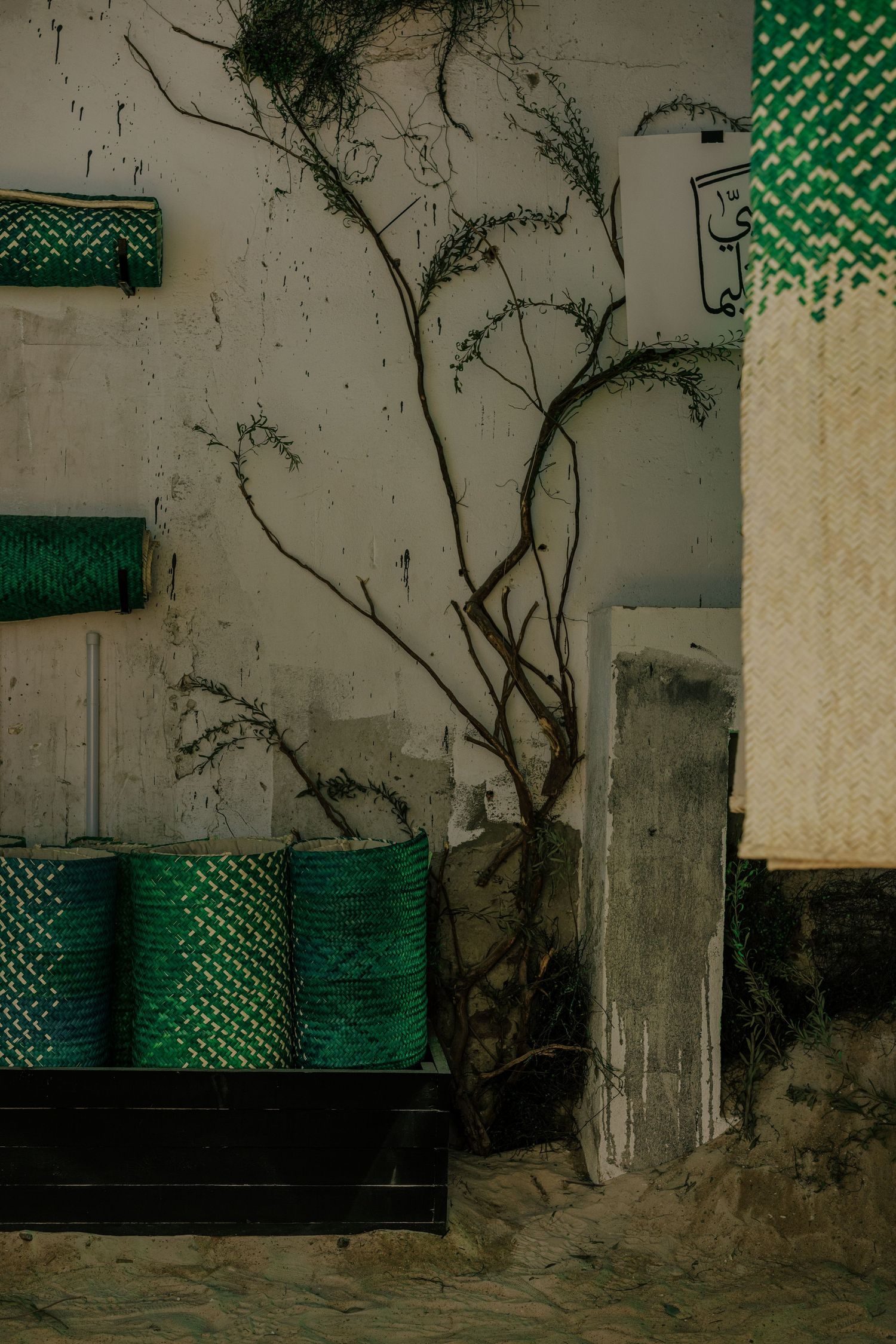 Green wicker baskets stacked against weathered white wall with bare branching tree in sandy setting.