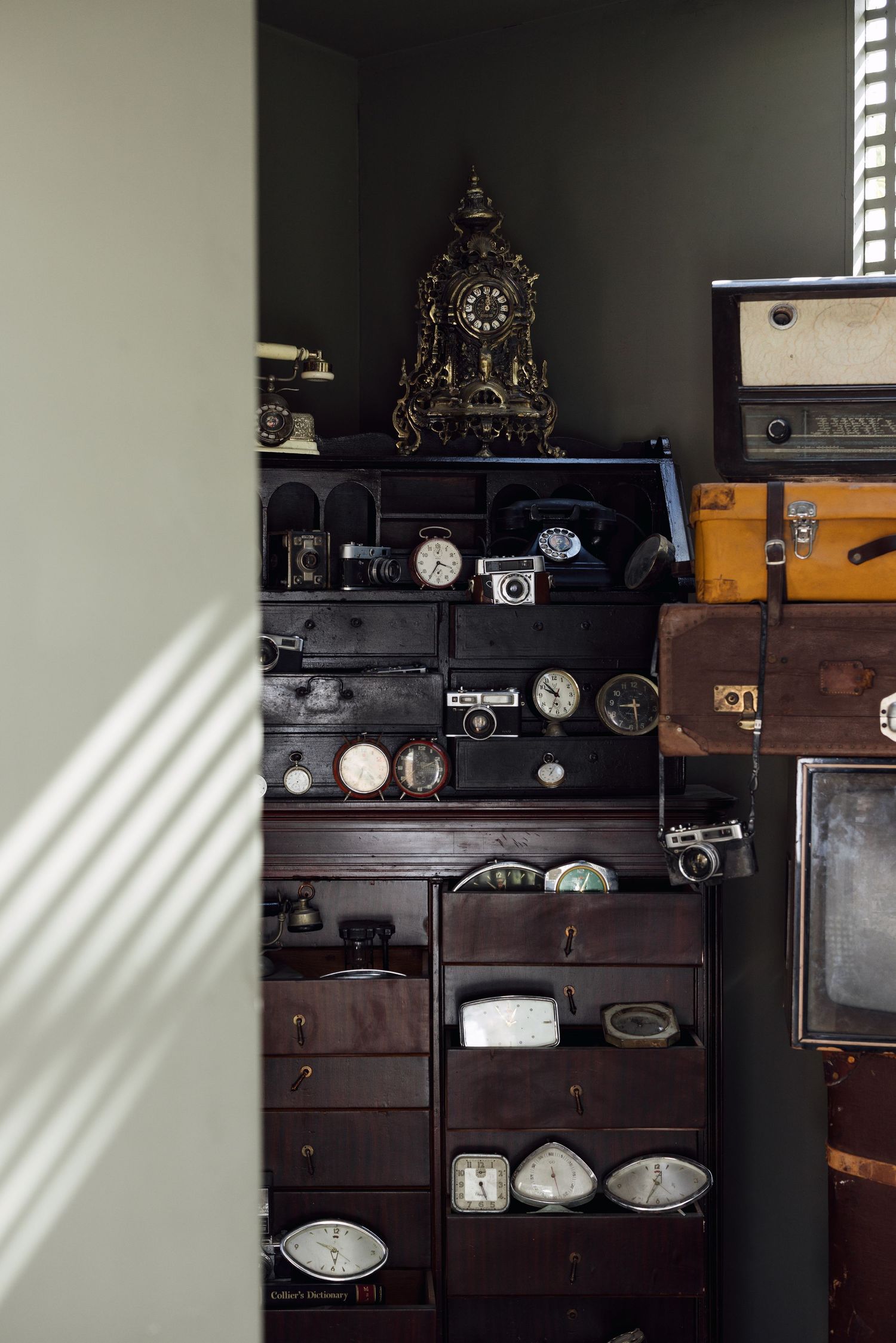 Antique wooden cabinet filled with vintage clocks, cameras, and collectibles in dimly lit room.