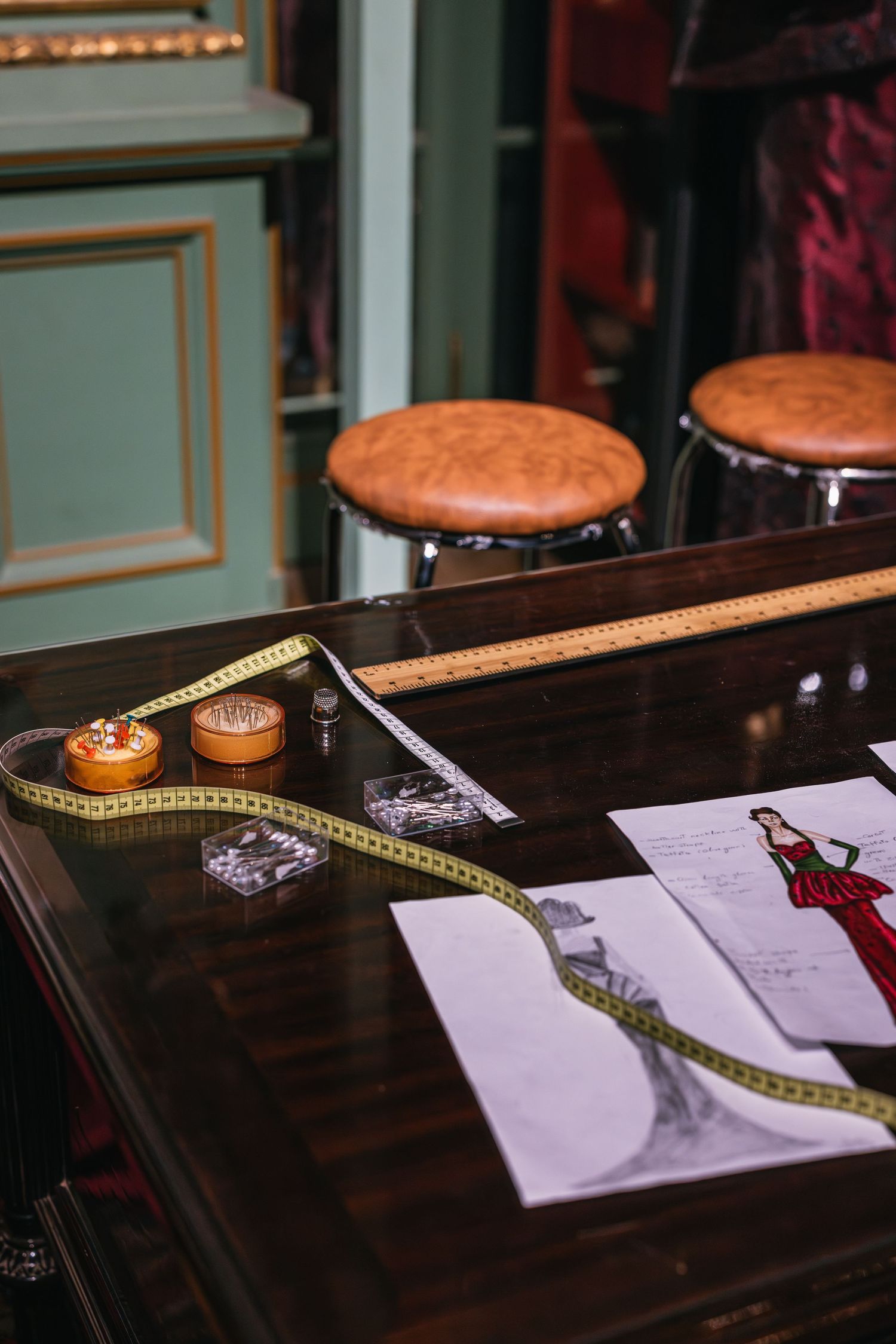 Fashion atelier table with dress sketches, measuring tape, pin cushions, and leather bar stools.