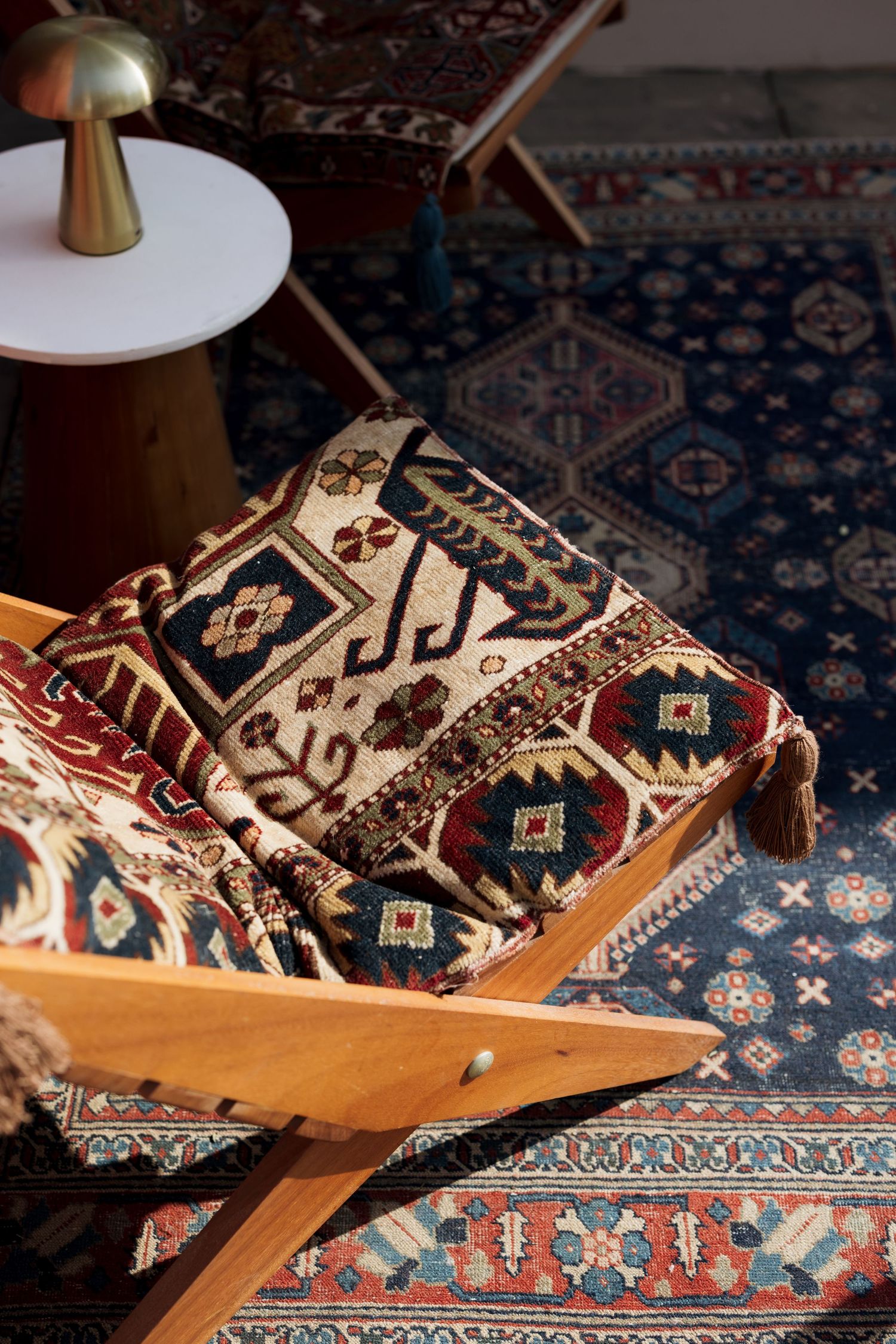 Folded traditional kilim rug with tassels on a wooden stand atop a blue Persian carpet with a brass lamp nearby.