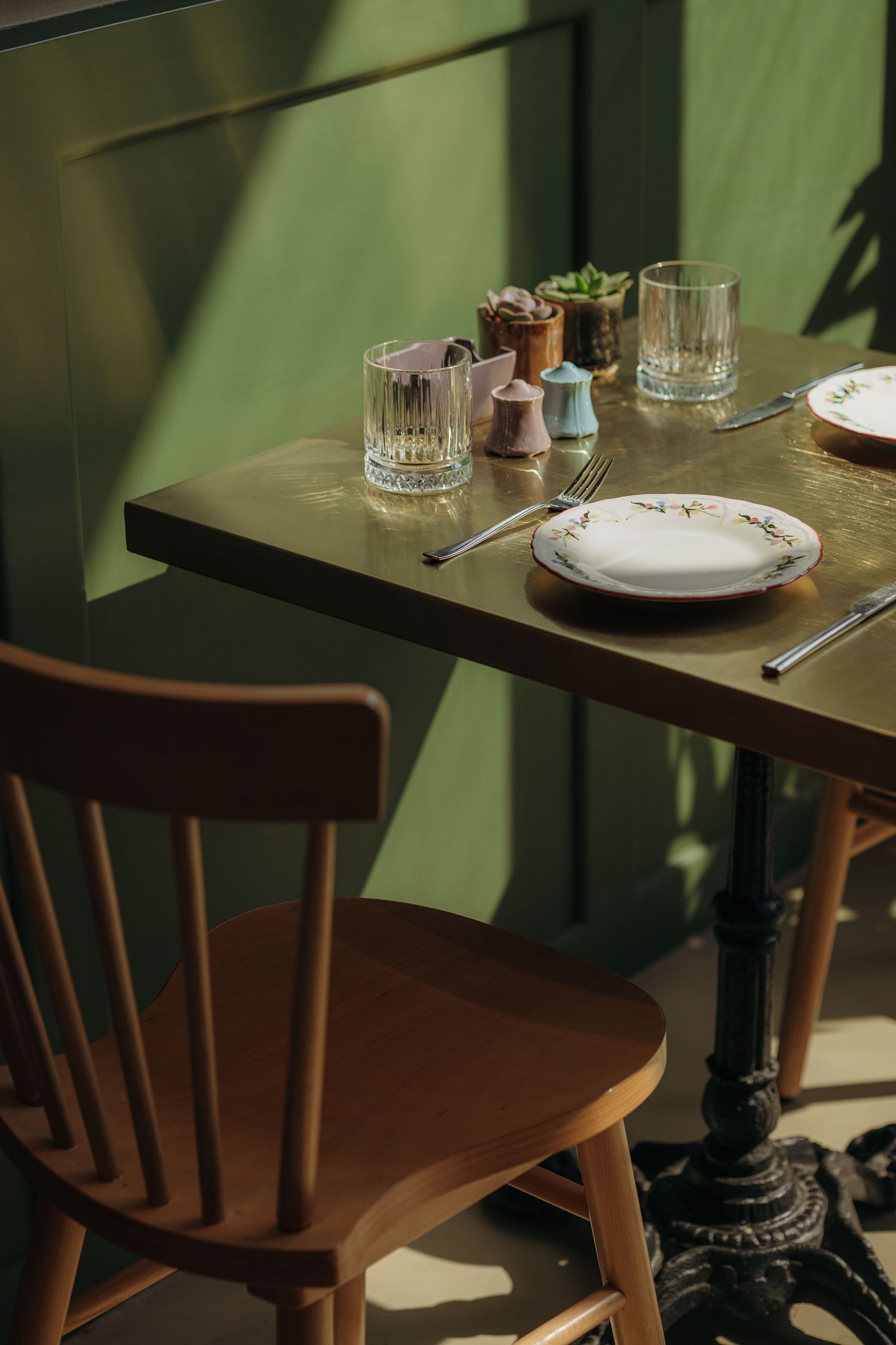 Sunlit restaurant table set with crystal glasses, white plate, and colorful salt shakers against a green wall.