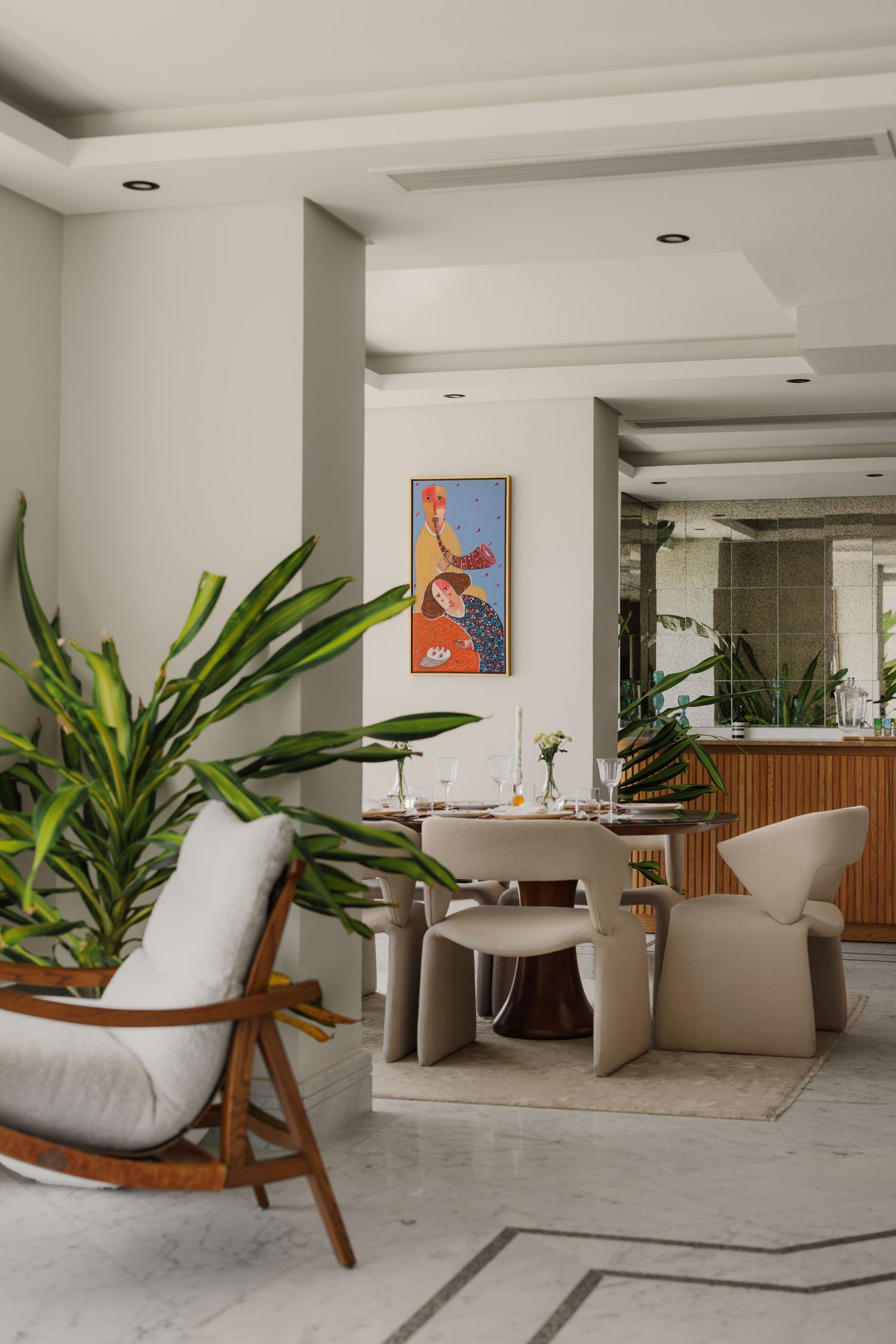 Contemporary open-plan dining area with tropical plants, sculptural chairs, colorful wall art, and neutral tones.