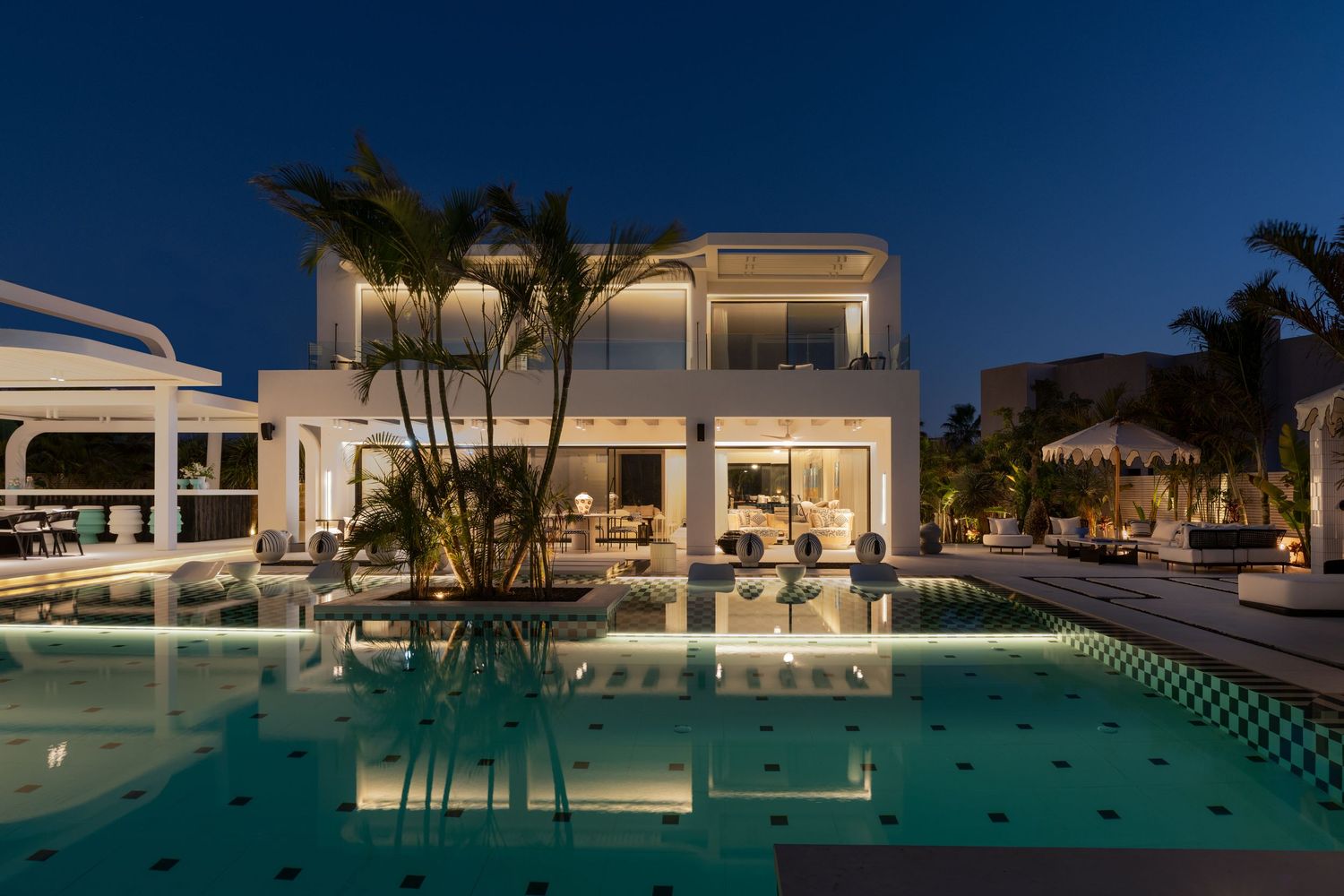 Spectacular white luxury villa reflecting in a glowing pool at night, framed by tropical palm trees and elegant architecture.