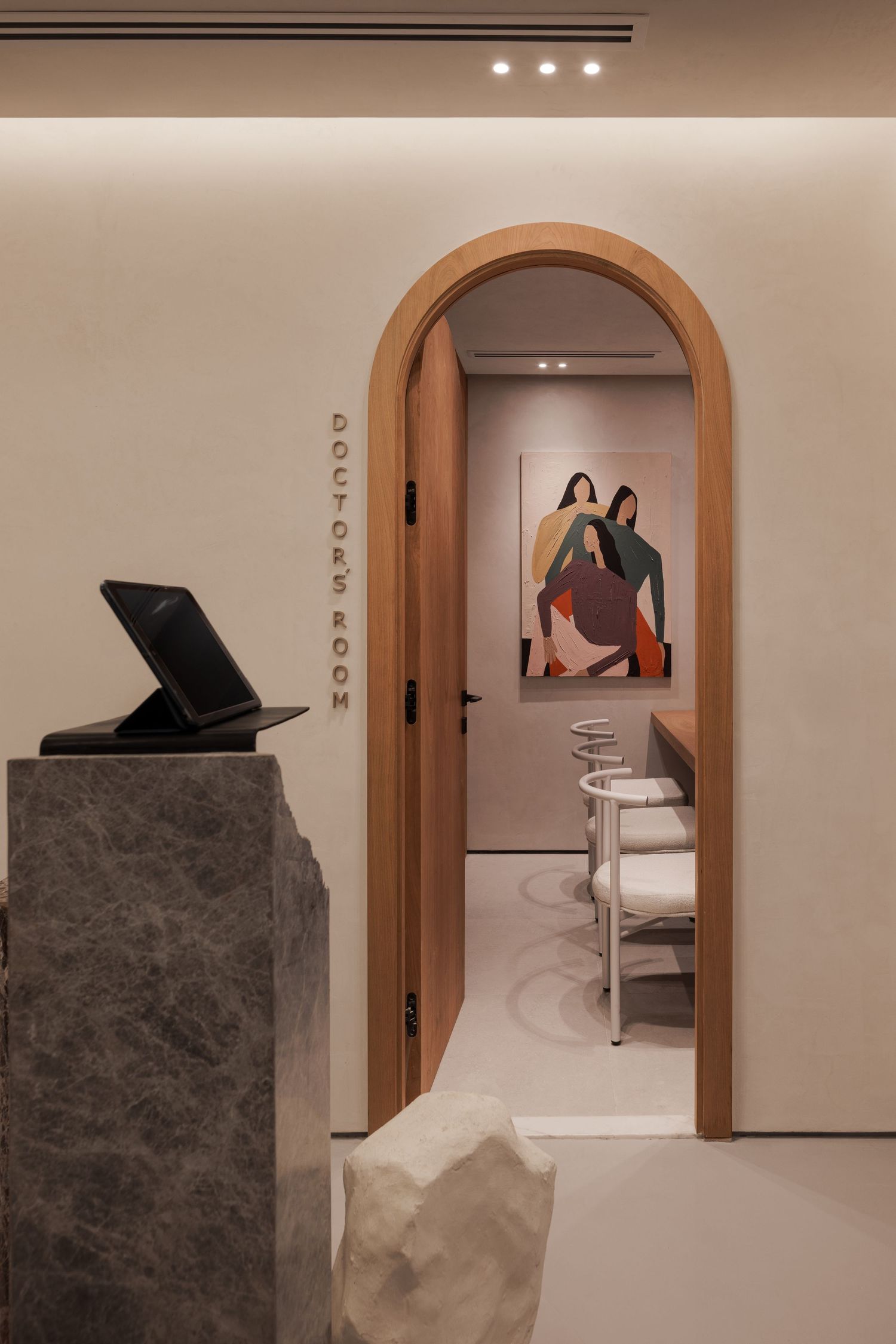 Minimalist boutique fitting room entrance with warm arched wooden door, abstract figurative artwork, and stone reception desk.
