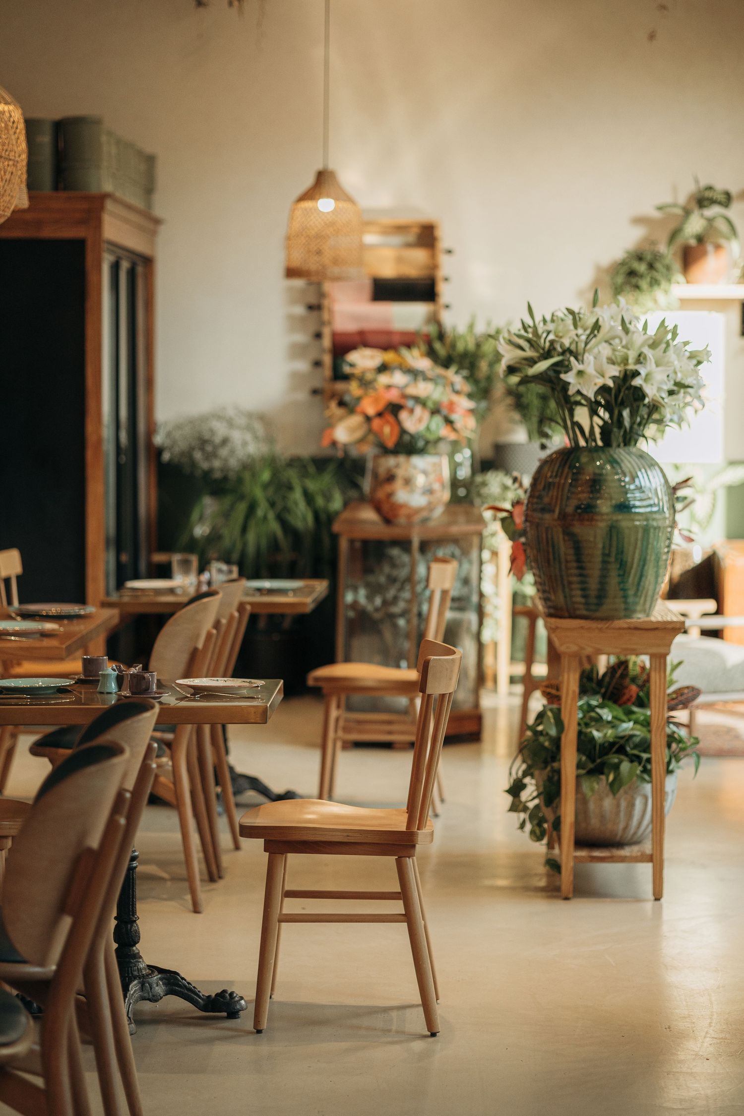 Bright cafe interior with wooden chairs, large decorative vases, fresh flowers, and lush green plants throughout.