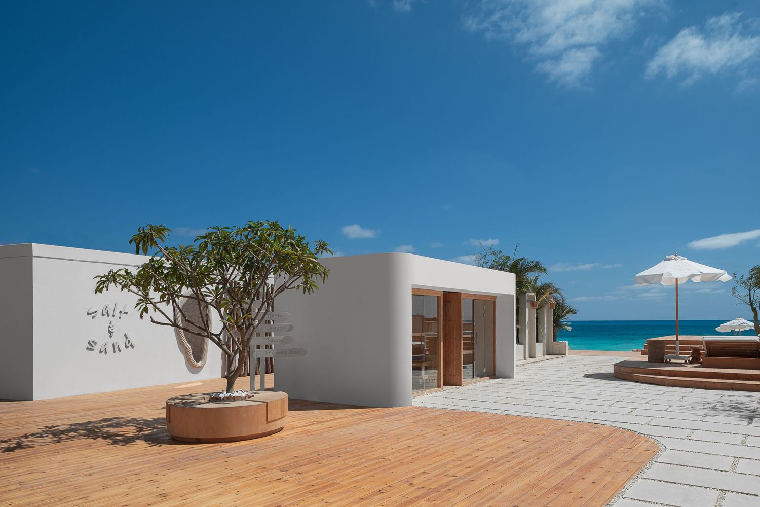 Minimalist white beachfront spa building with tropical tree, wooden deck, and stunning turquoise ocean backdrop.