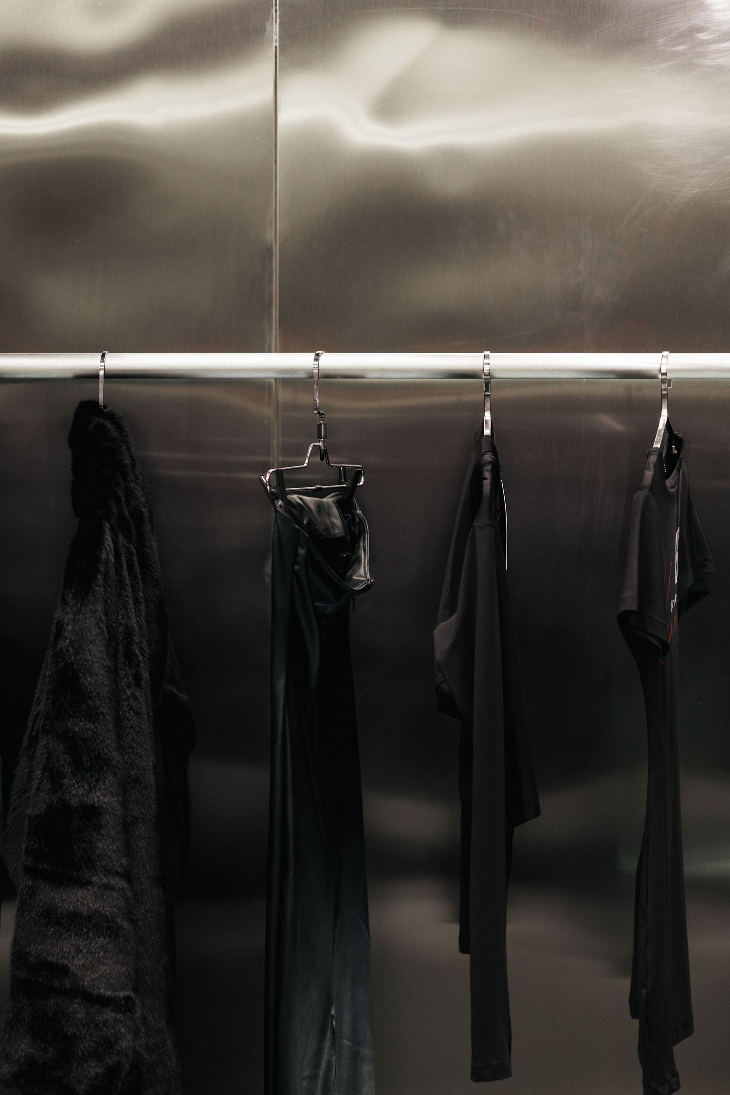 Black garments hanging on a chrome rail against a dark metallic wave-textured wall in a high-end fashion store.