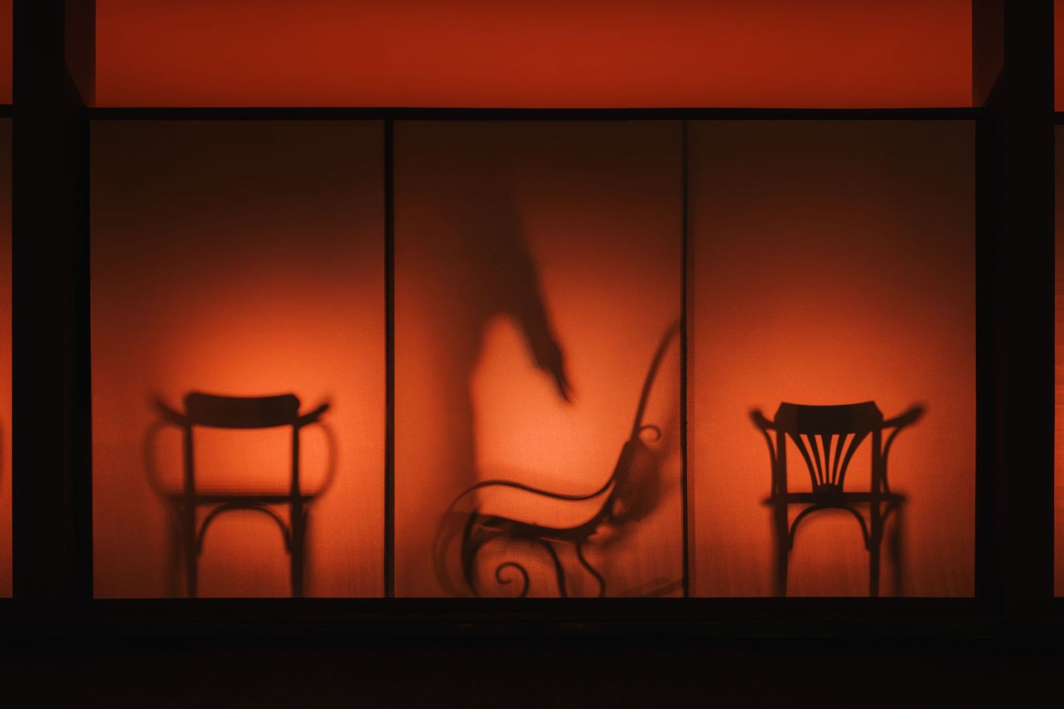 Dramatic red-lit window display showing silhouettes of ornate wooden chairs against glowing orange backdrop.