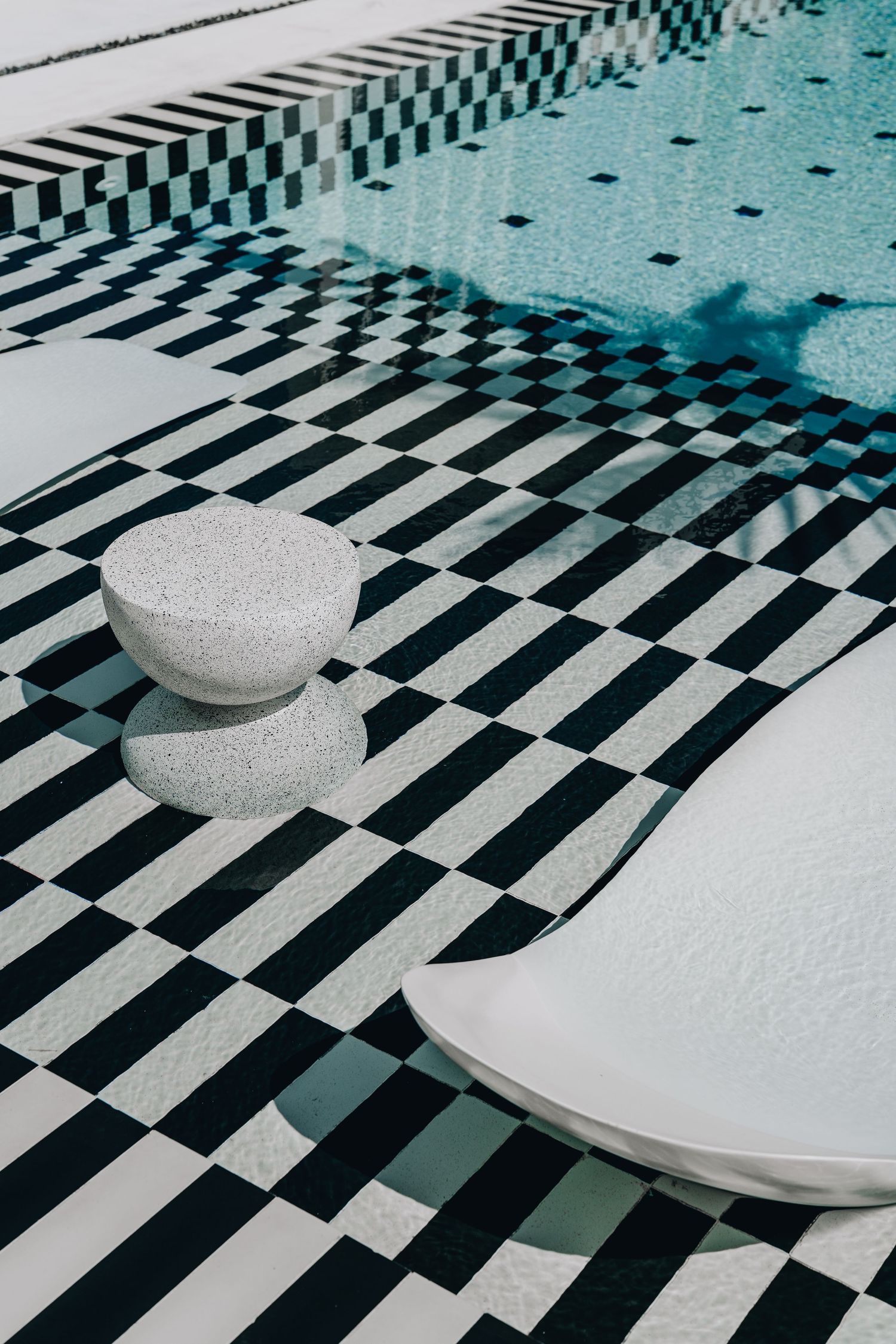 Black and white checkered pool deck with stacked stone sculpture and white lounger beside a turquoise swimming pool.