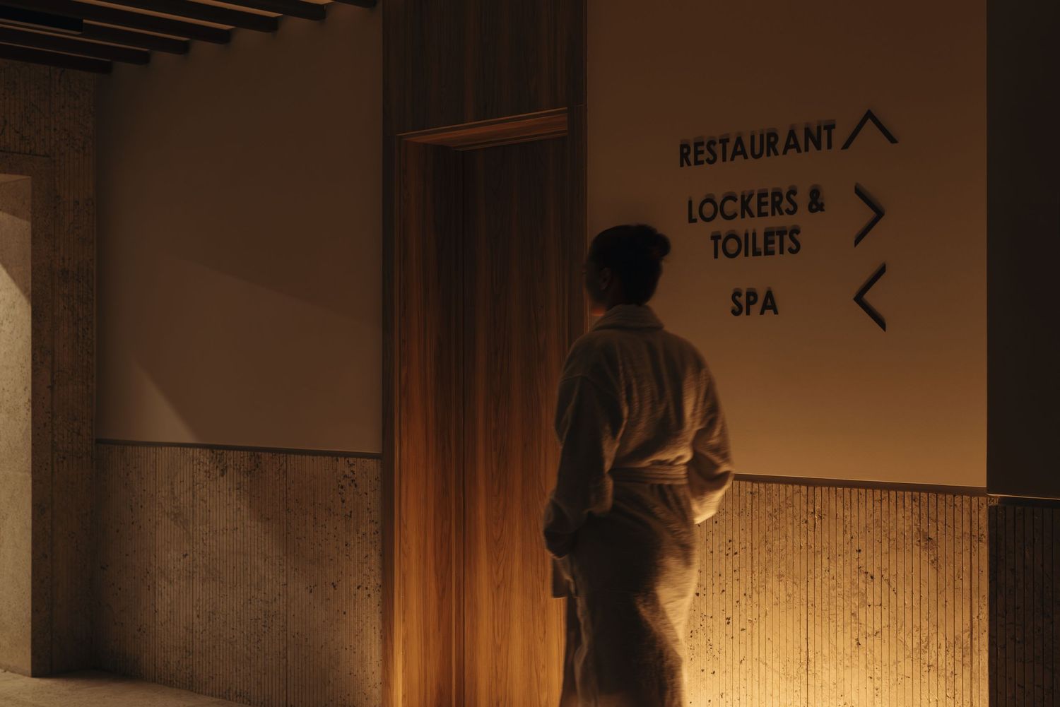 Person standing before illuminated directional signs pointing to restaurant, lockers, toilets, and spa.