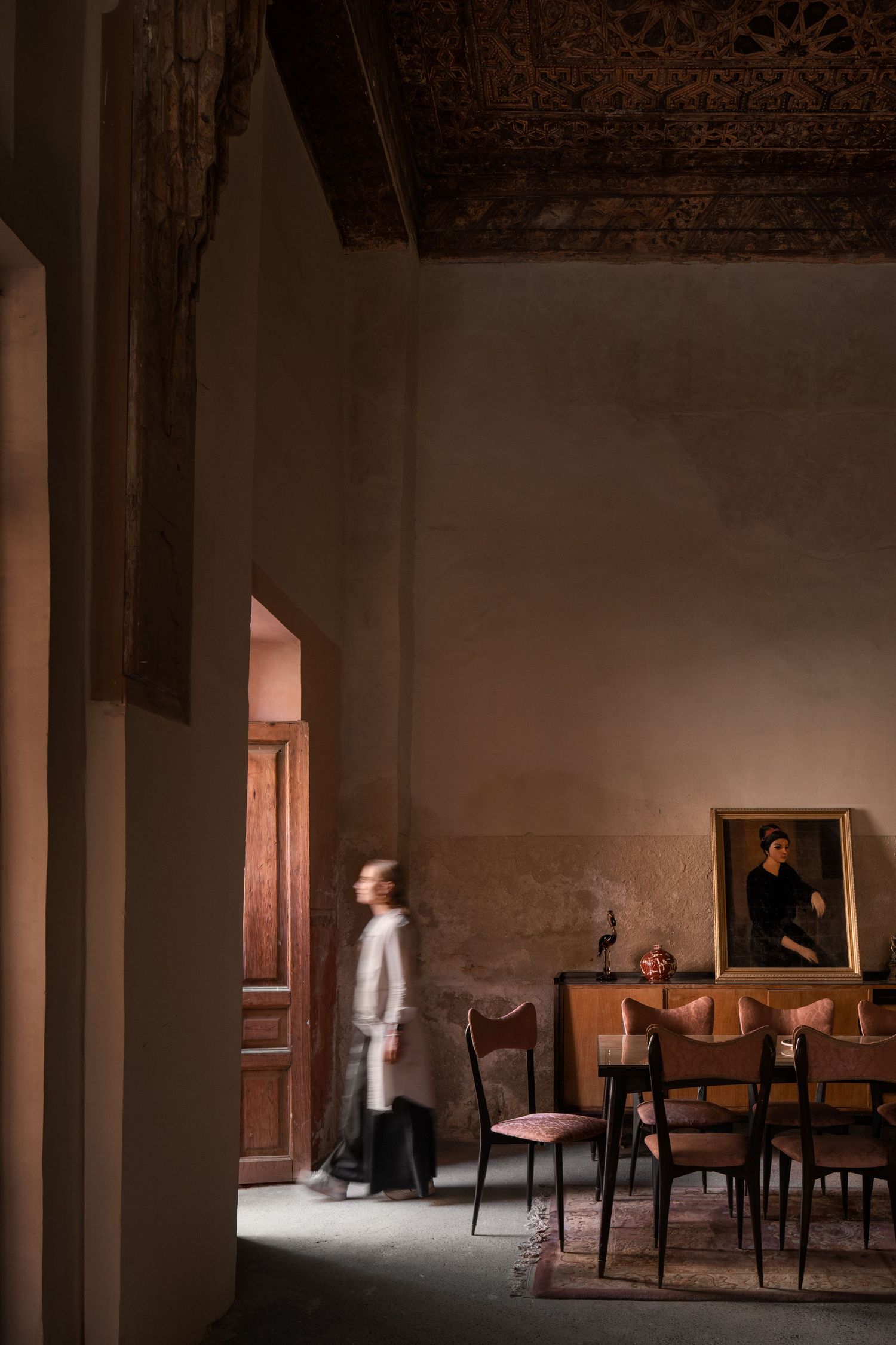 Moody Moroccan riad dining room with motion-blurred figure, carved wooden ceiling, leather chairs, and framed portrait.