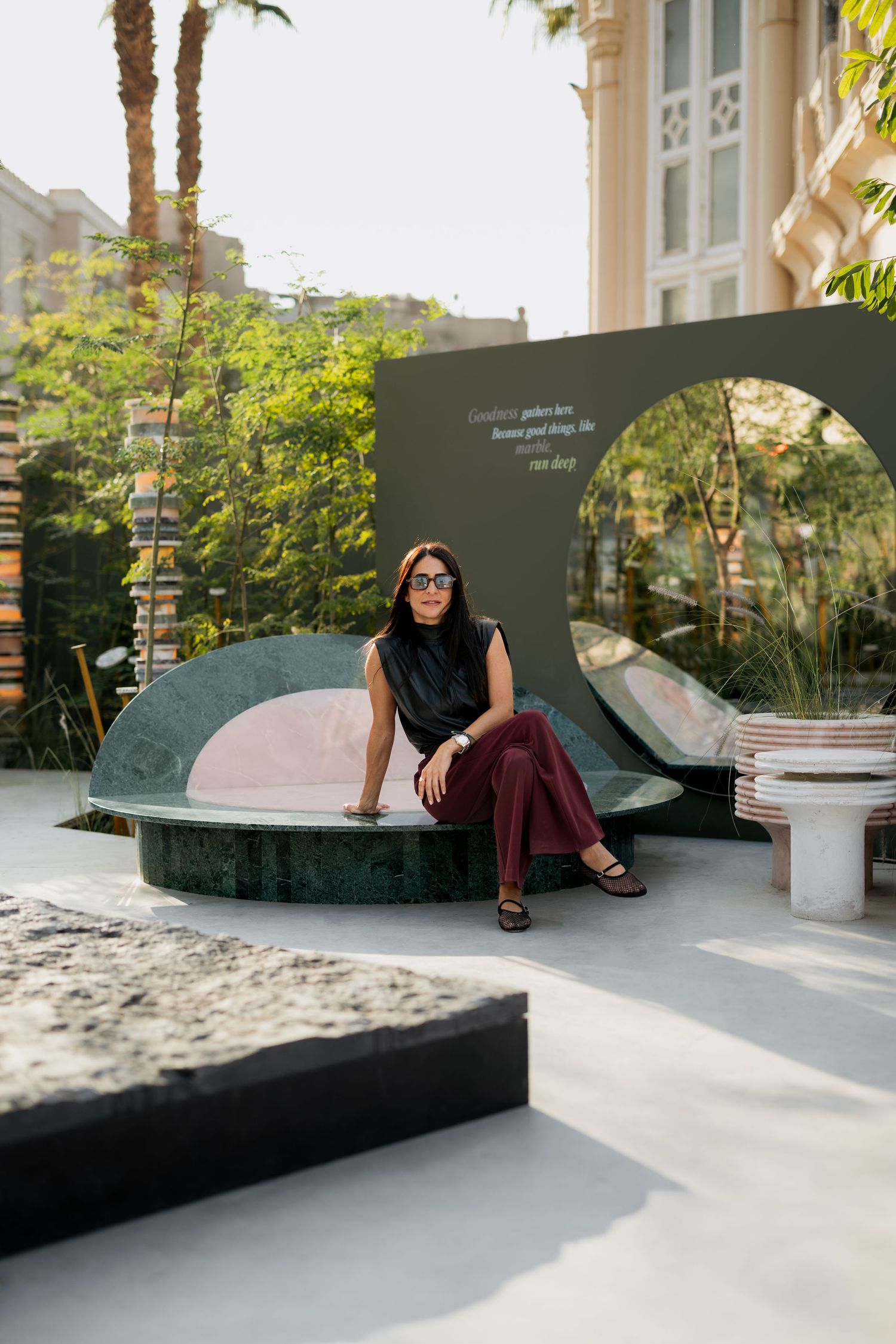 Woman in sunglasses sitting on a green marble curved bench installation in a lush outdoor garden.