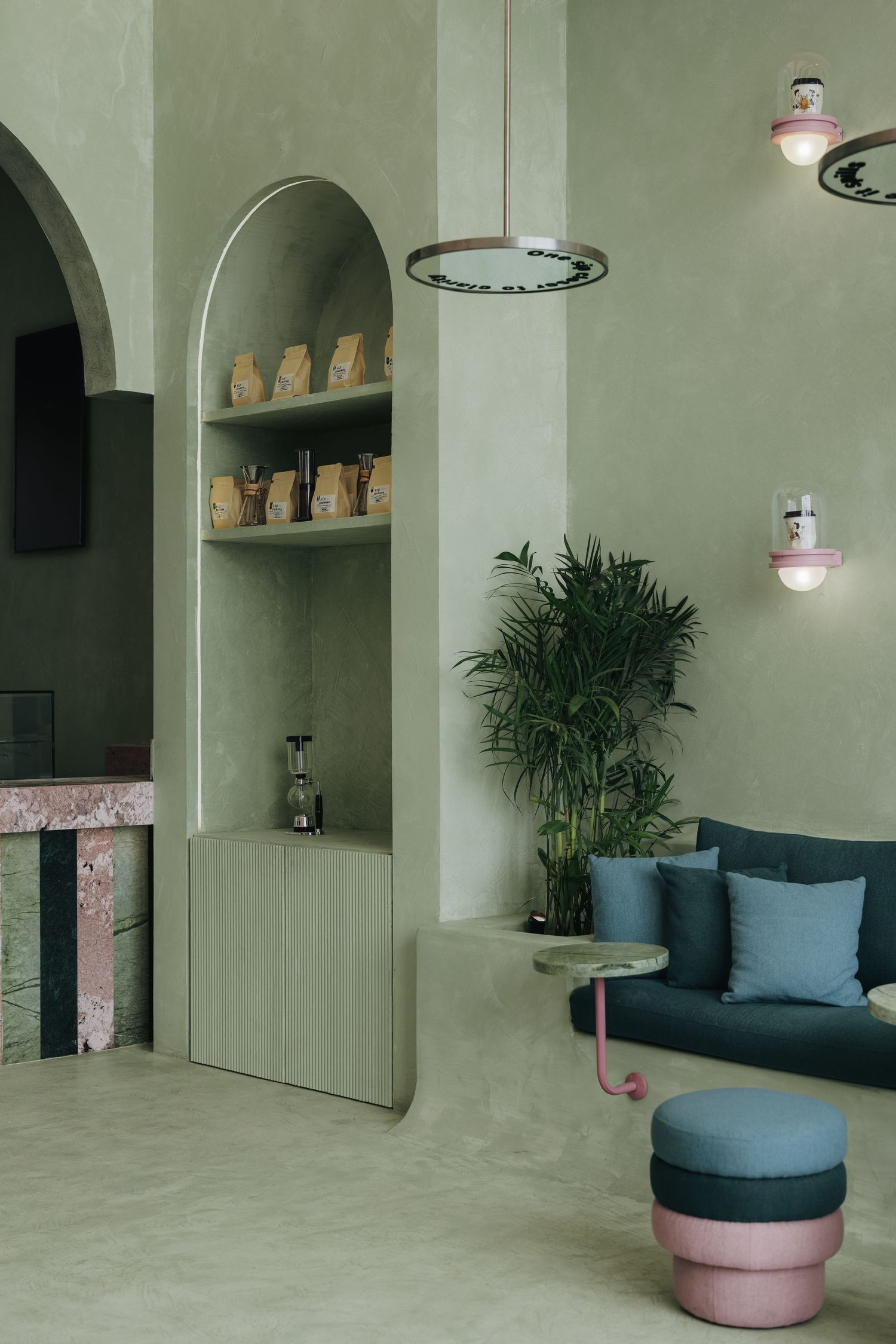 Sage green cafe interior with arched shelving, teal sofa, pink ottomans, and decorative pendant lights.