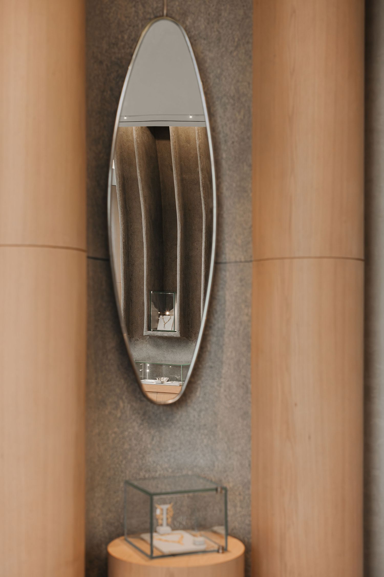 Oval wall mirror reflecting jewelry displays, flanked by warm wood columns above a glass showcase pedestal.