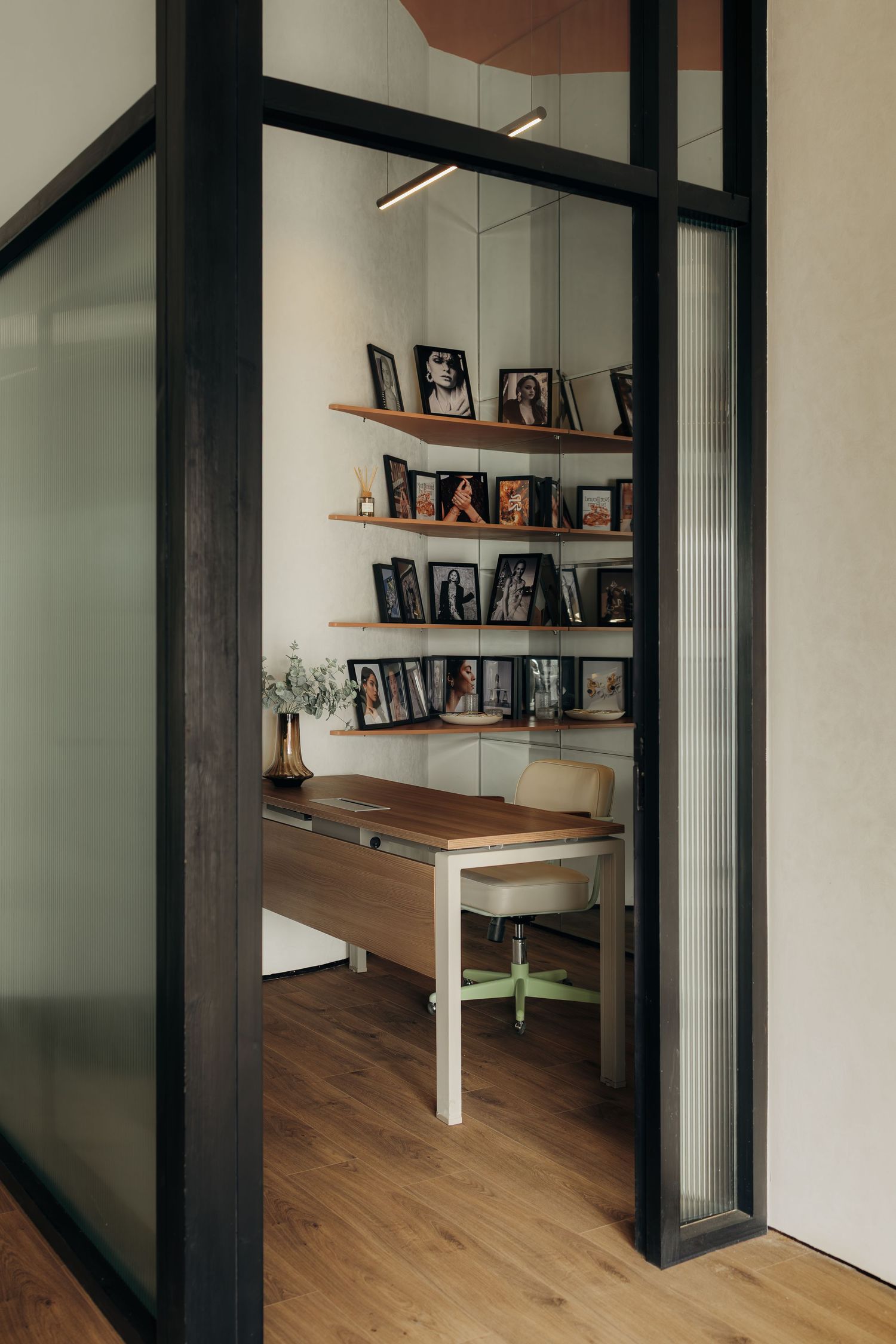 Small private office viewed through black-framed glass doors with wood desk, shelving, and framed photographs.