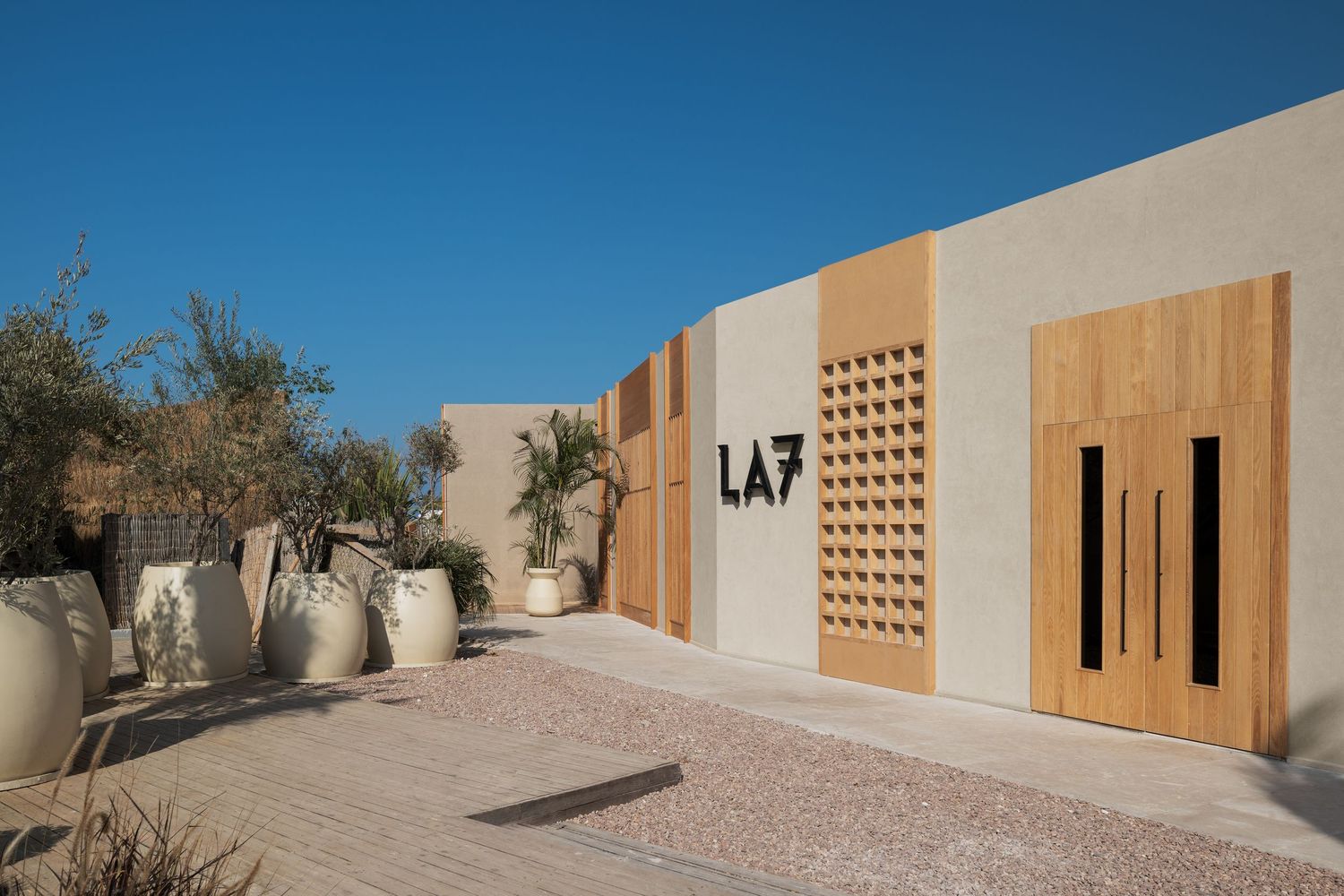 LA7 luxury boutique entrance with warm beige walls, wooden doors, olive trees, and decorative ceramic pots.