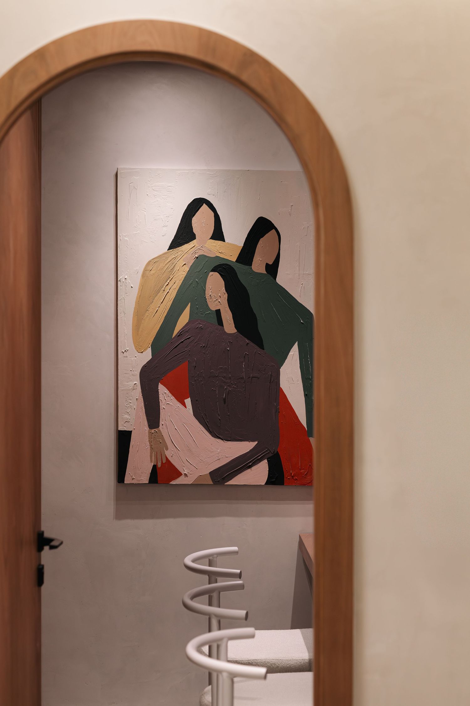 Arched wooden doorway framing abstract figurative painting of three women in earthy tones above white wire bar stools.