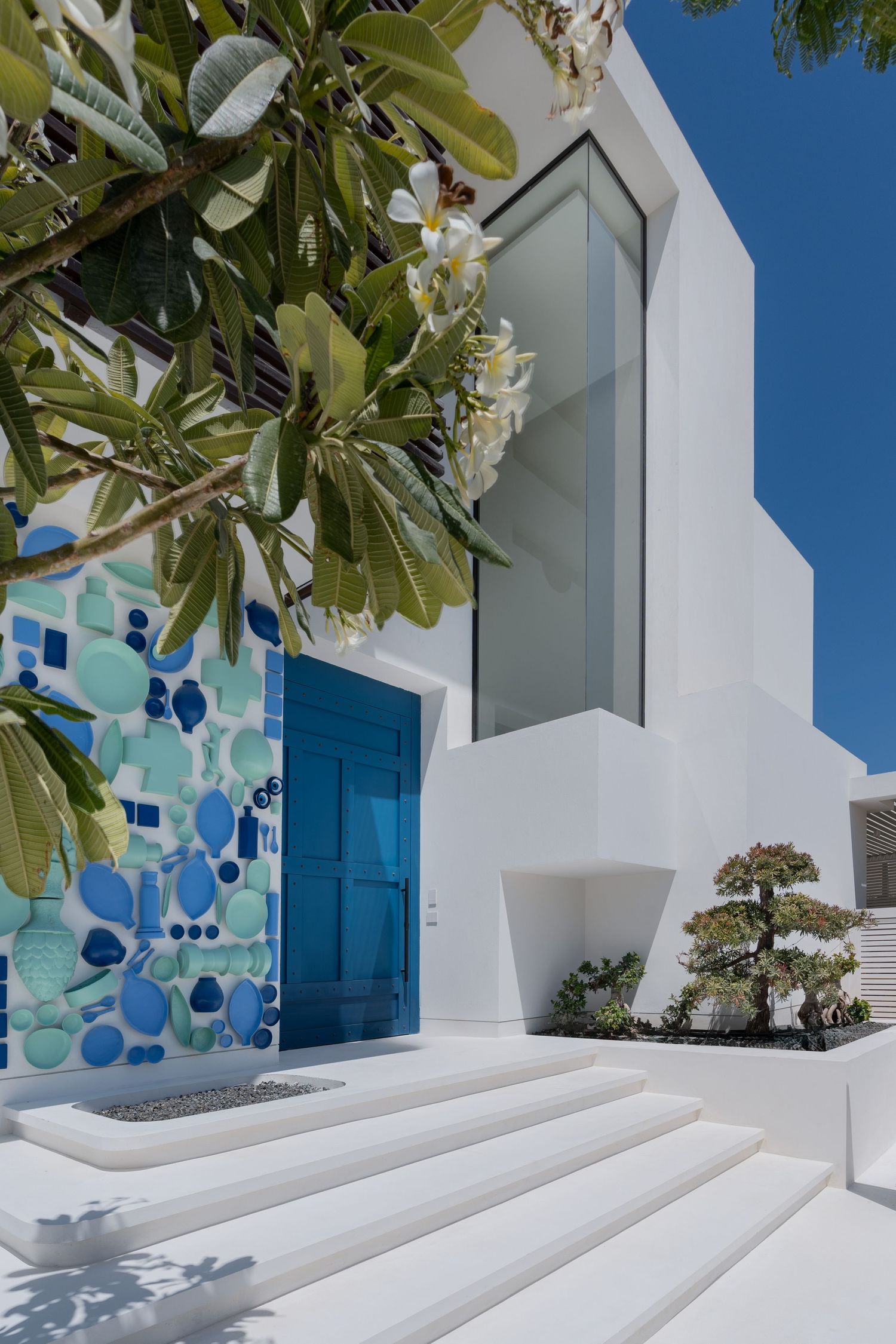 Modern white villa entrance with vibrant blue mosaic tiled door surround, glass window, and tropical frangipani tree.
