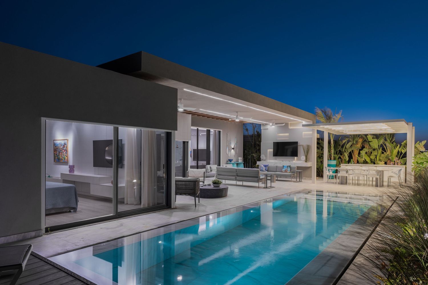 Luxury villa exterior at night featuring illuminated pool, open living space, and pergola bar area.