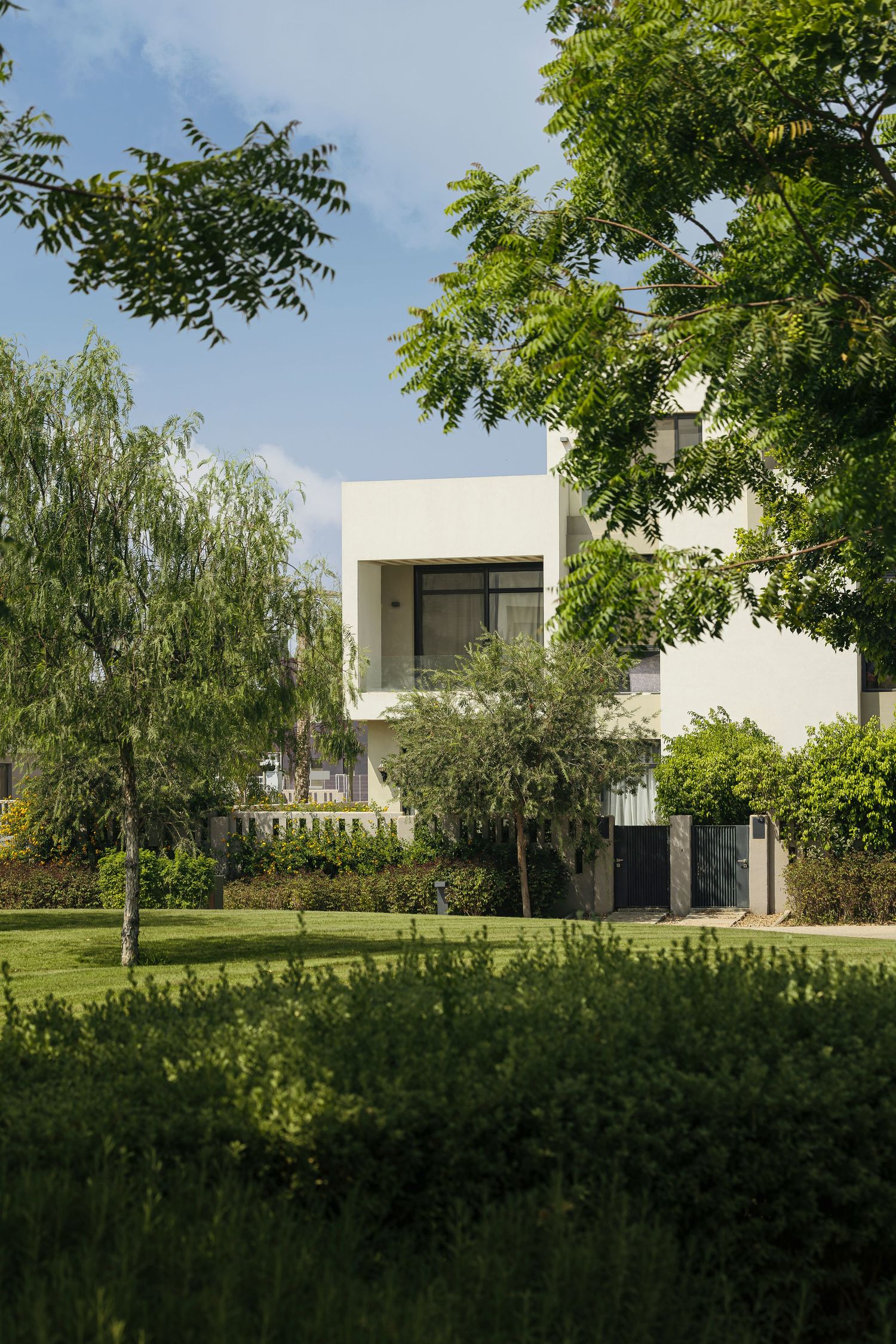 Modern white two-story villa surrounded by lush green trees and manicured lawn under a blue sky.