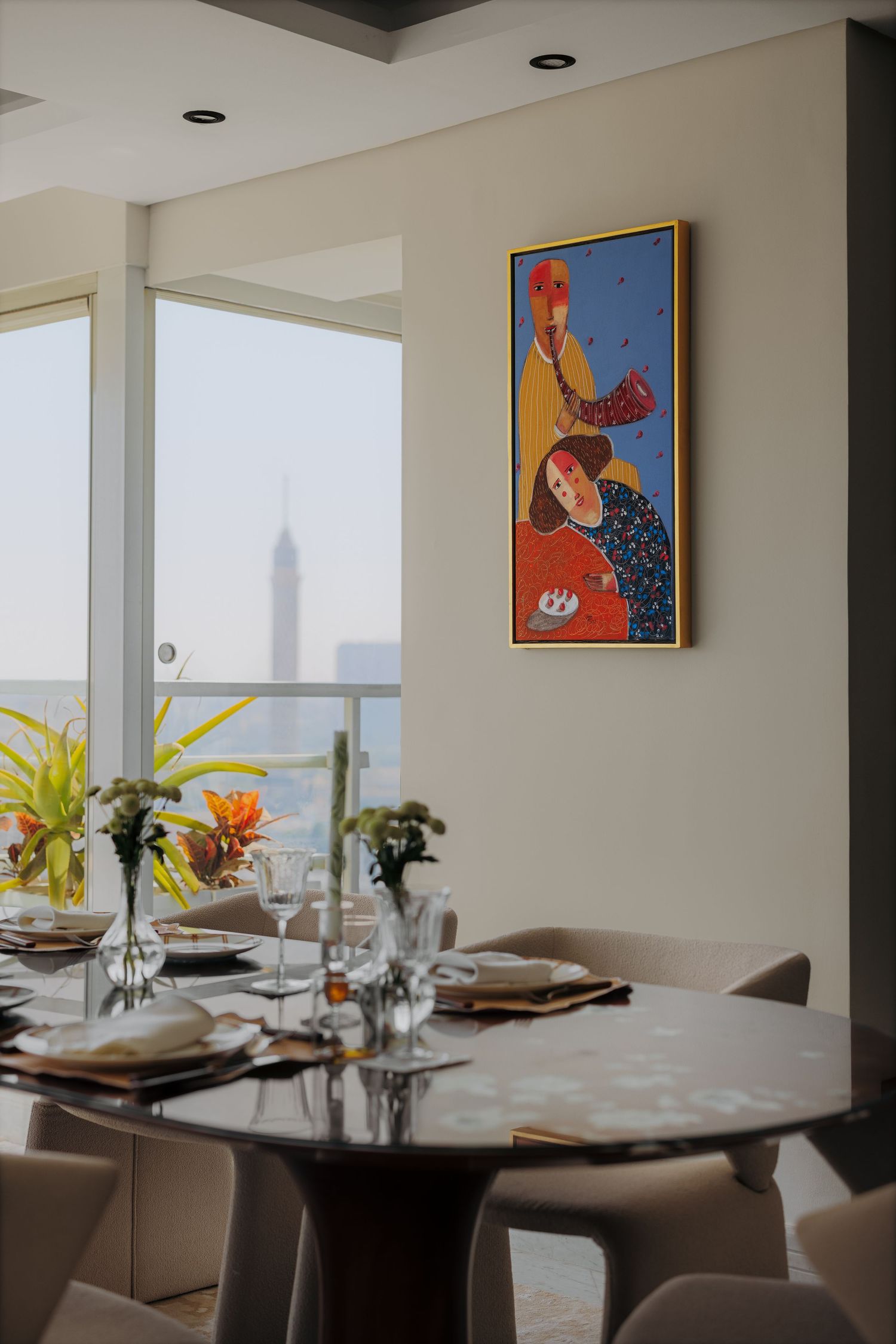 Modern dining room with round glass table, colorful artwork, and Cairo Tower visible through large windows.