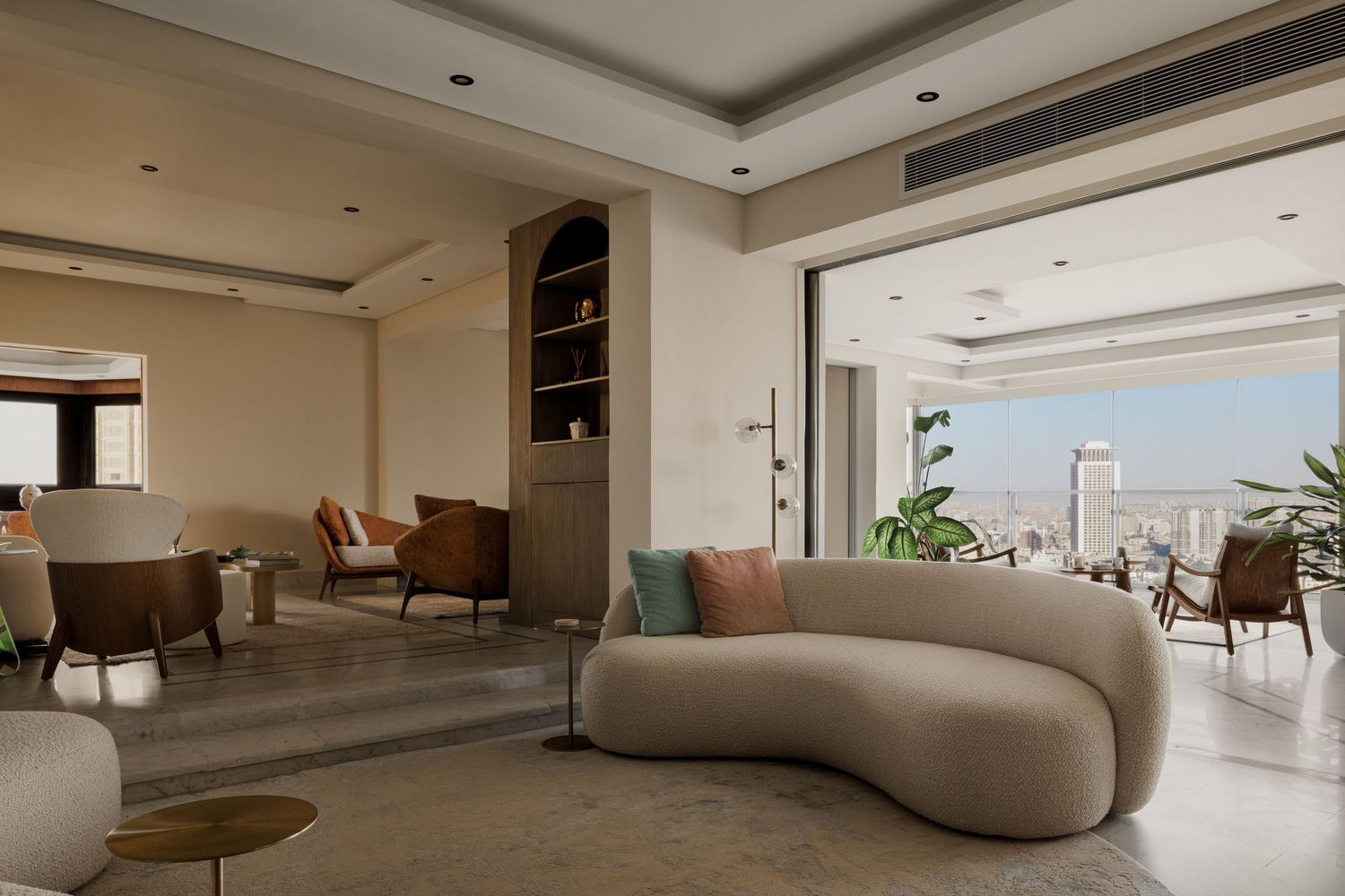Modern open-plan living room with curved cream sofa, panoramic city views, and warm neutral contemporary furnishings.