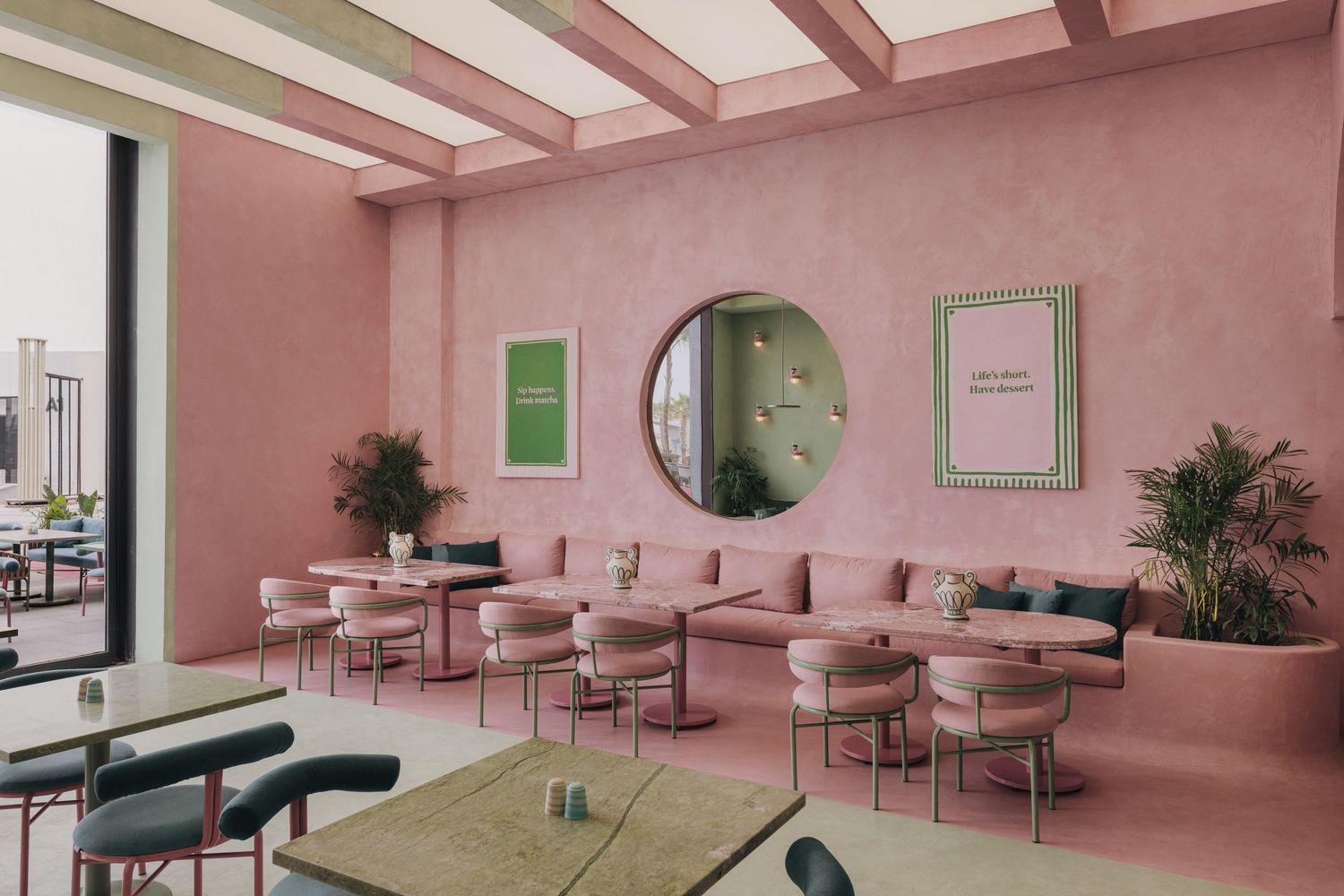 Pink minimalist restaurant interior with round mirror, green framed art, and matching pink chairs.