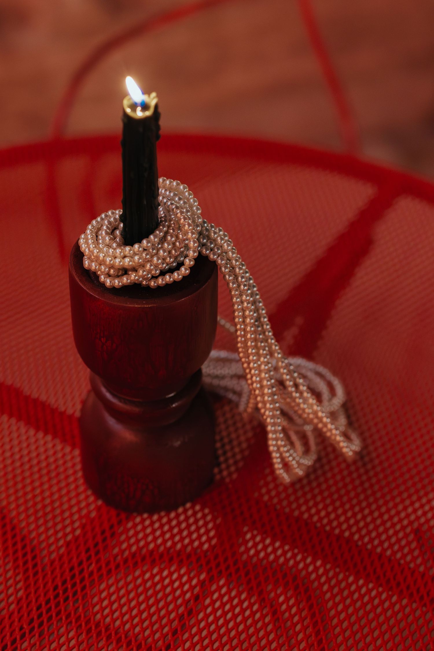 Black candle burning in a dark wooden holder wrapped with silver beaded necklace on a red mesh table.