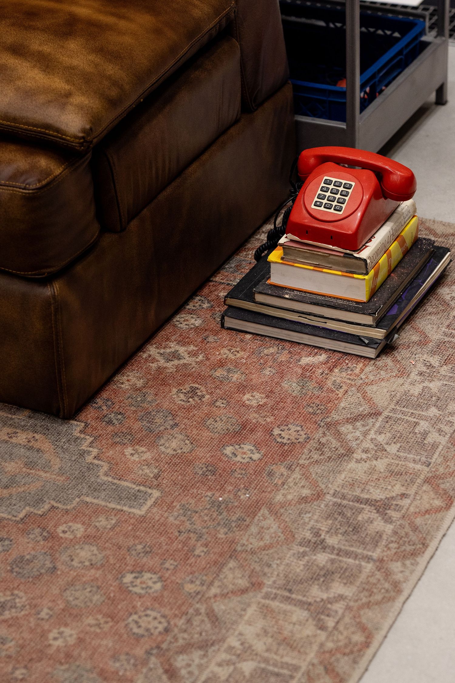 Vintage red rotary phone resting on stacked books beside a brown leather couch on a patterned rug.
