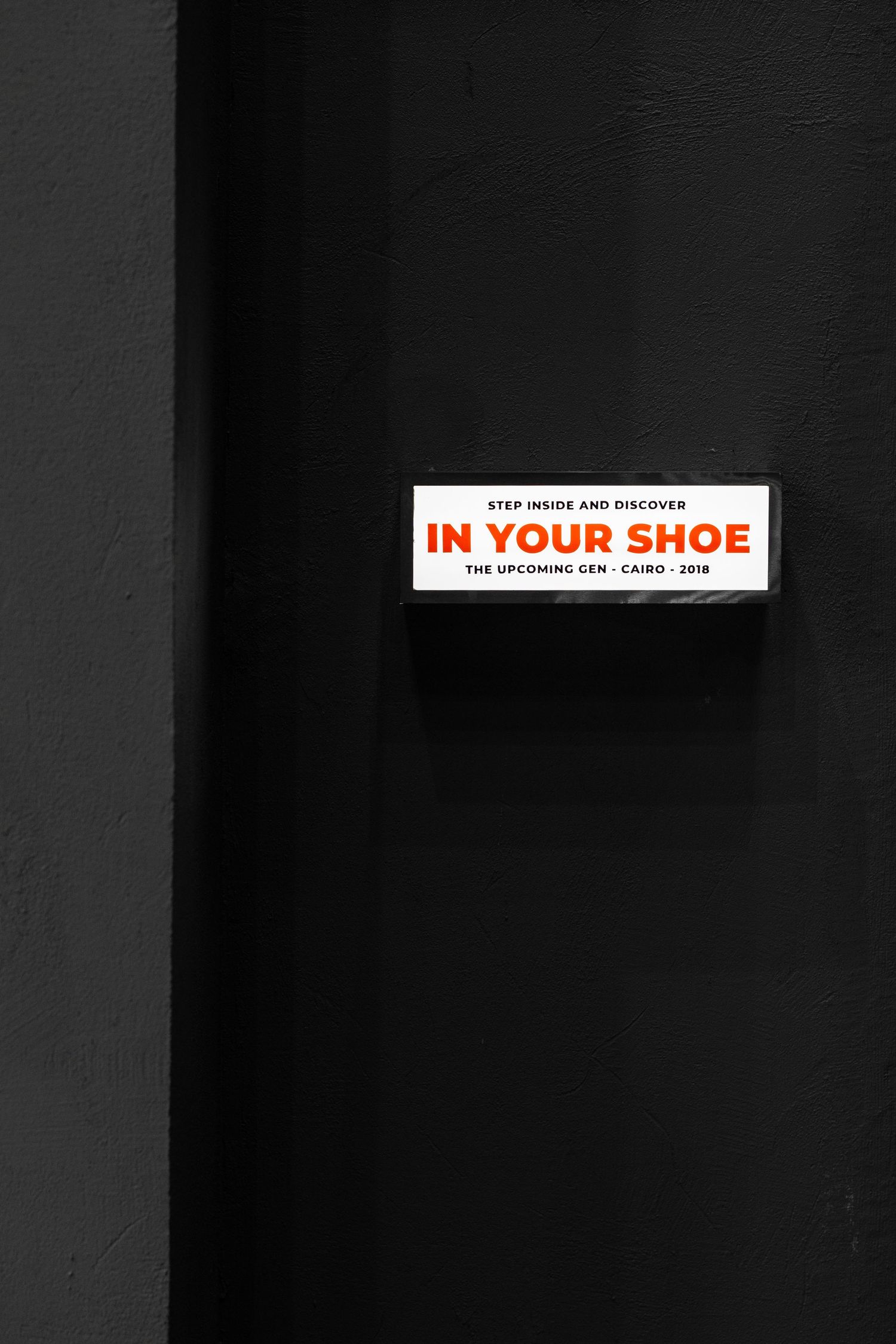 Small white rectangular sign reading In Your Shoe on dark black wall with red and black typography.
