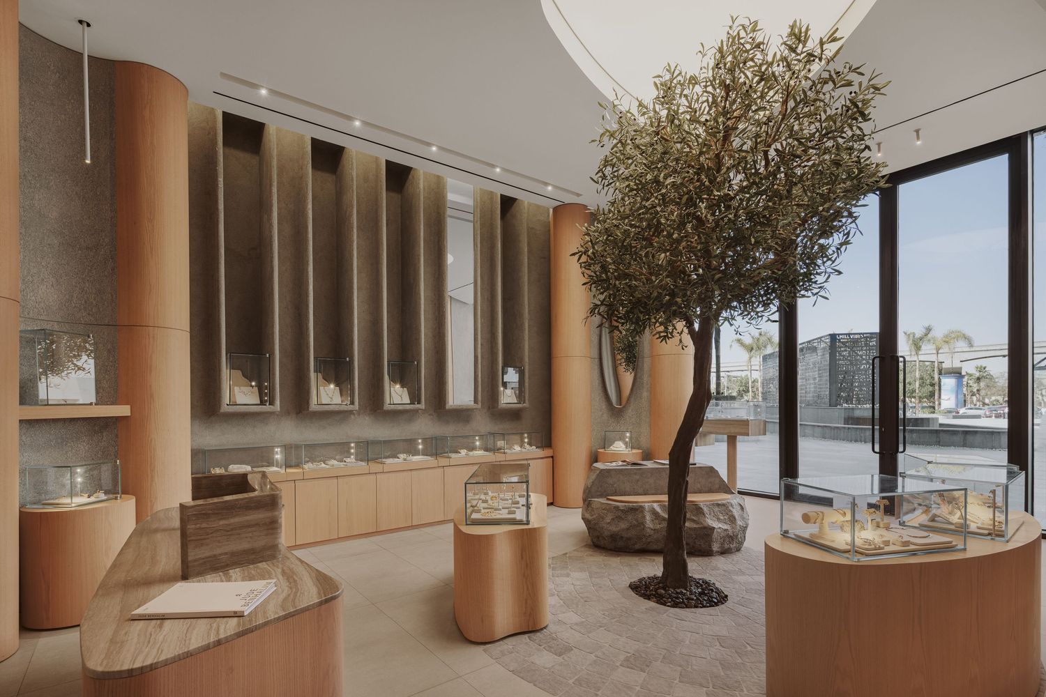 Upscale jewelry store interior with curved wood columns, olive tree centerpiece, and illuminated display pedestals.