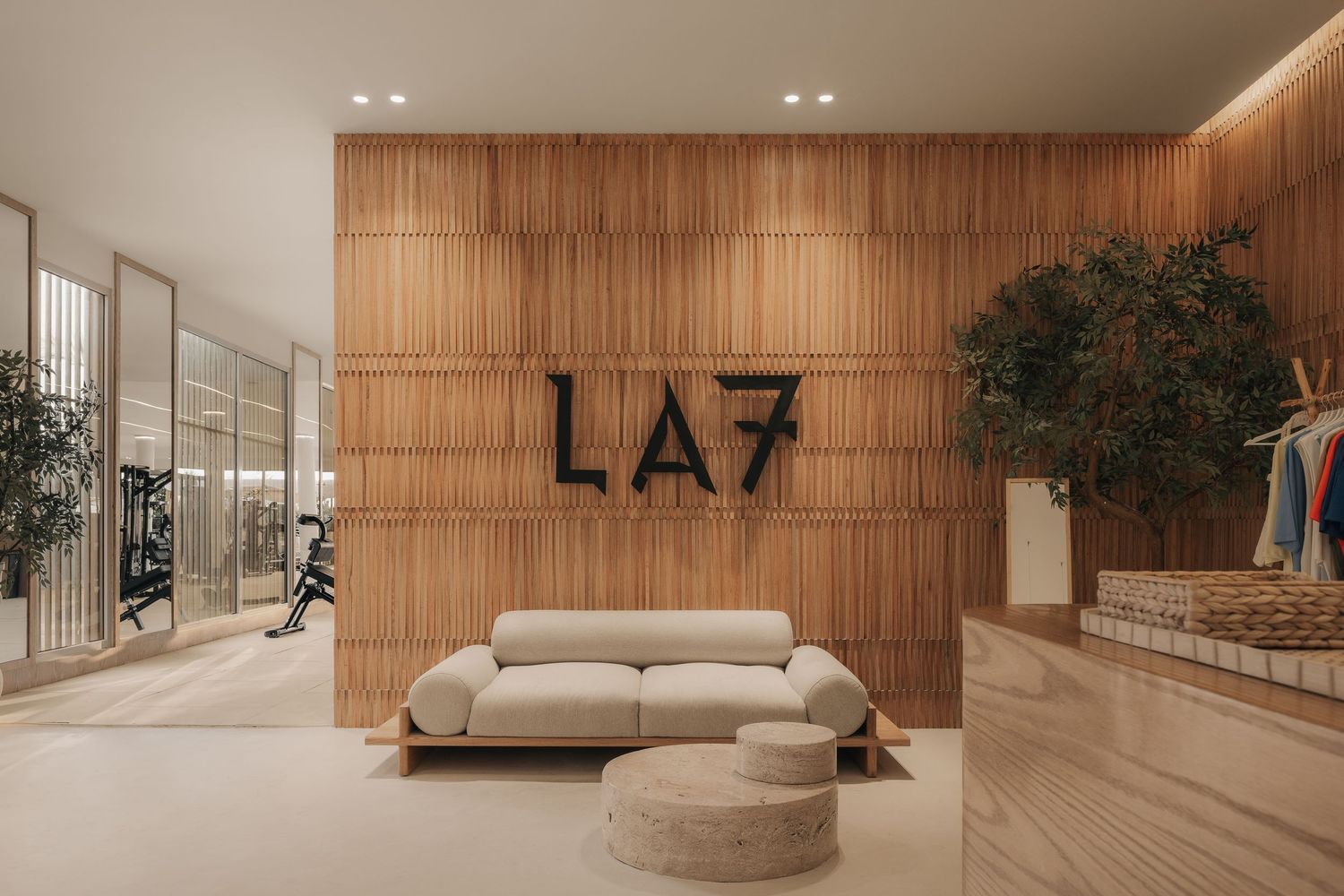 Modern LA7 branded lobby featuring warm wood slat walls, minimalist white sofa, ottoman, and indoor trees in neutral tones.