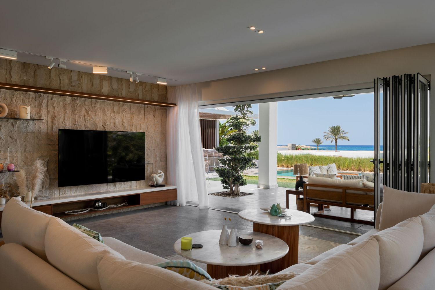 Elegant beachfront villa living room with cream sectional sofa, marble coffee table, and panoramic ocean views through open doors.