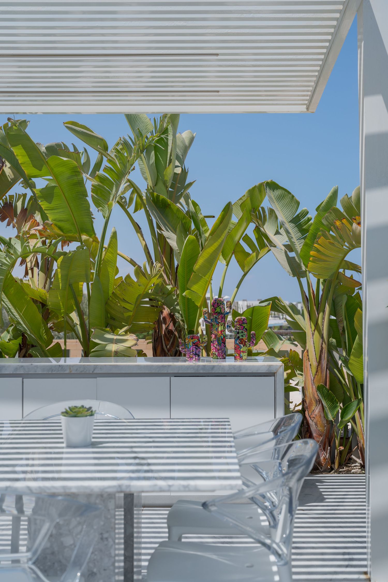 Outdoor marble dining table under white pergola surrounded by lush tropical banana plants in sunlight.