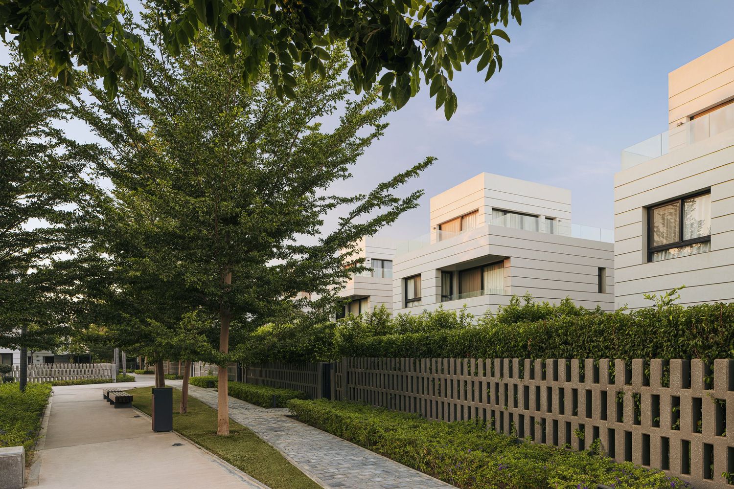 Modern residential buildings with white facades surrounded by lush green trees and manicured hedges along a paved walkway.
