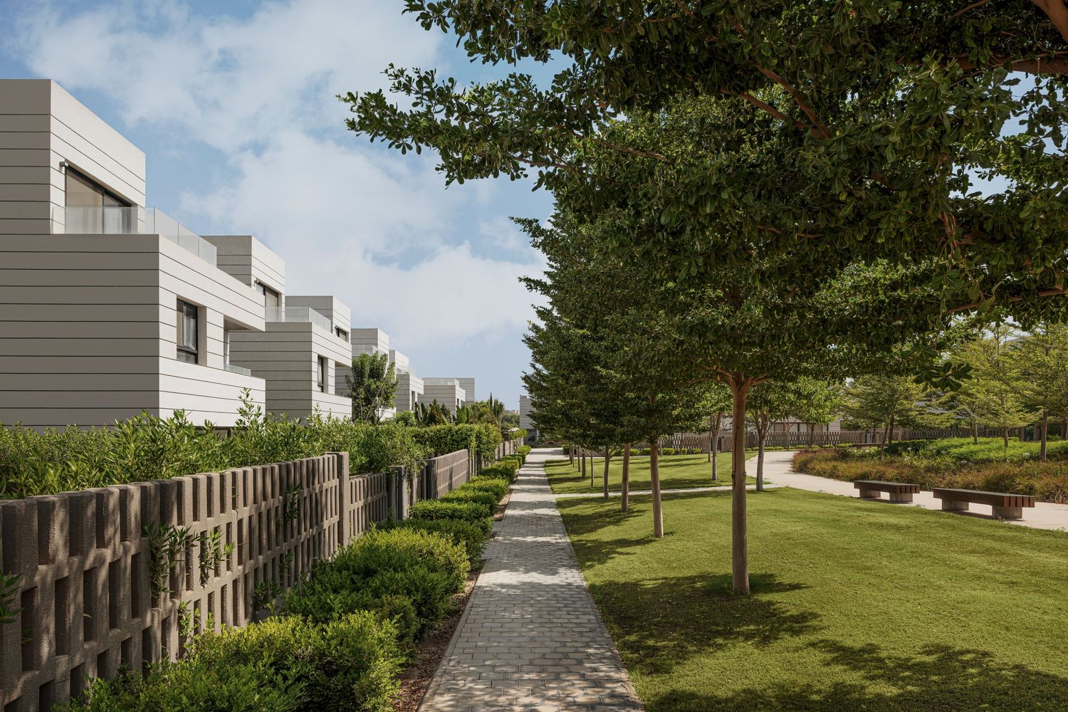 Modern residential buildings with lush green trees and manicured hedges lining a scenic cobblestone pathway on a sunny day.