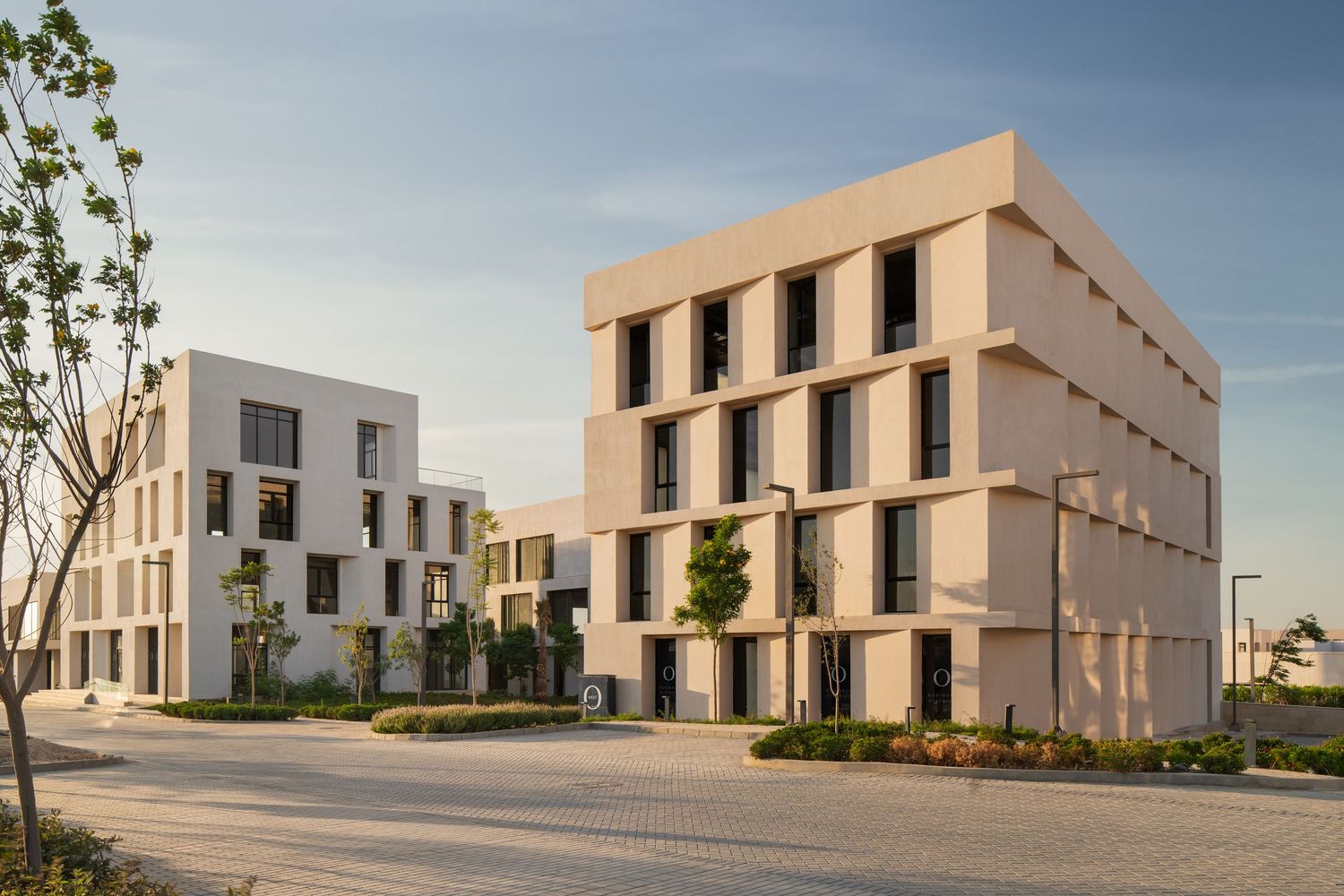 Modern minimalist beige residential buildings with clean geometric facades and young trees in a sunny setting.