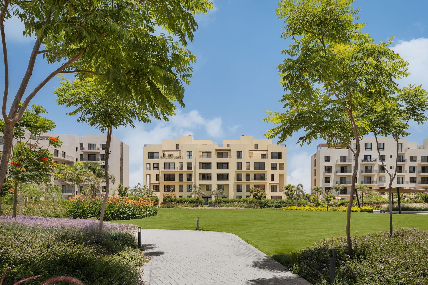 Scenic residential complex with manicured green pathways, mature trees, and beige apartment buildings in a gated community.