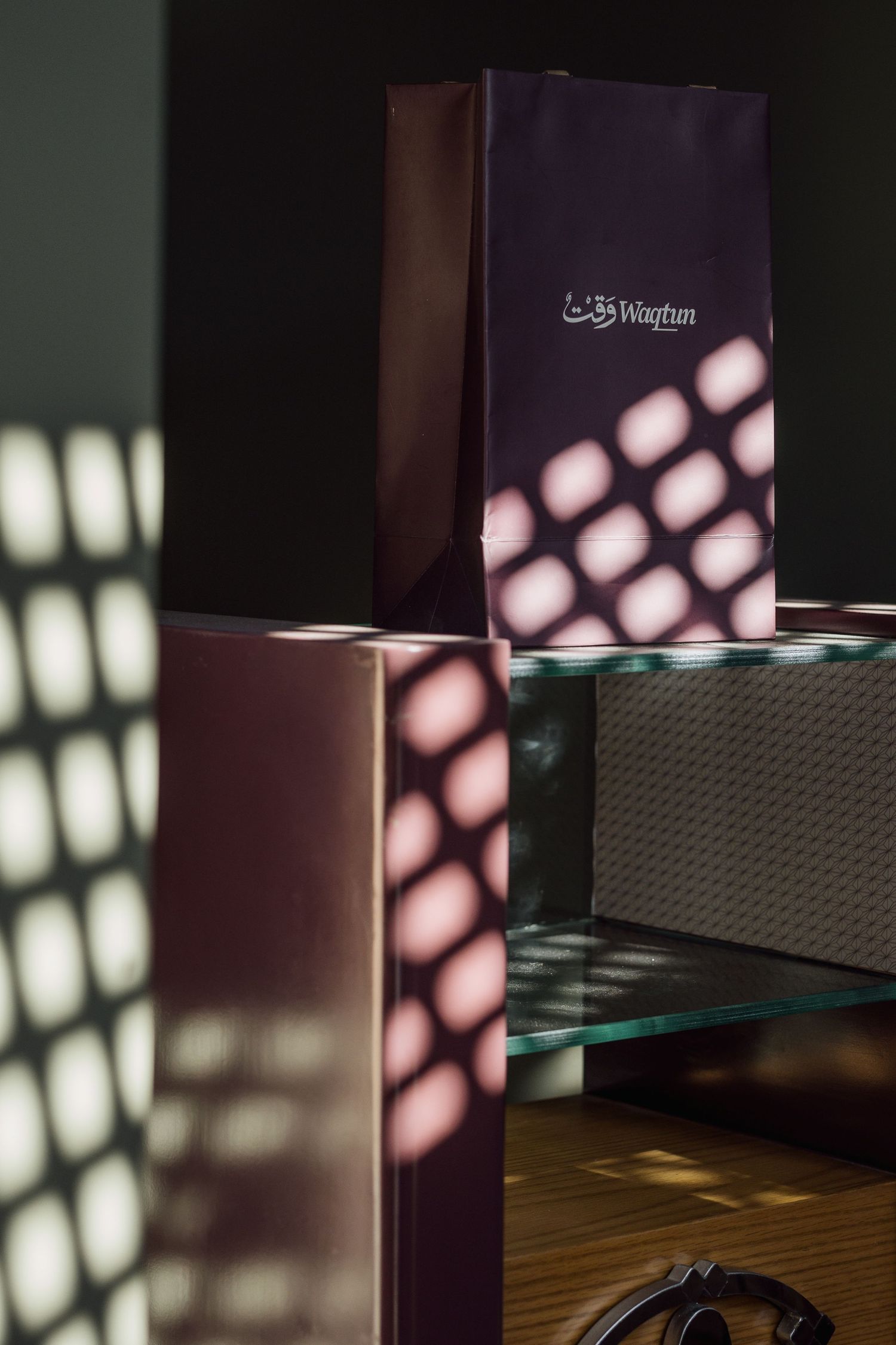 Purple Waqtun branded gift bag on glass shelf with geometric shadow patterns on dark wall.