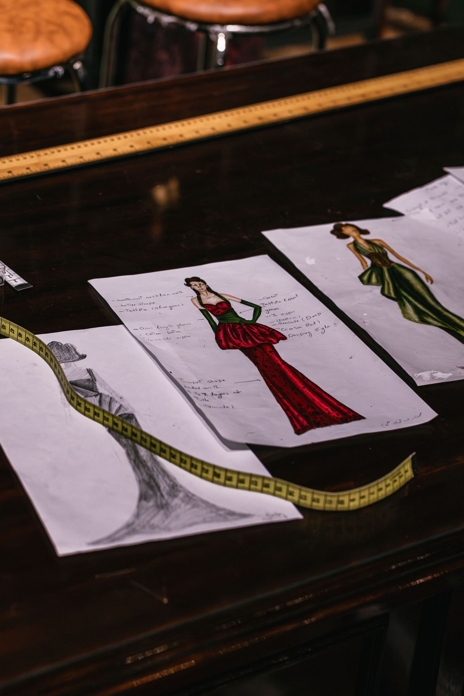 Fashion design sketches of elegant gowns laid on a dark wooden table with a yellow measuring tape.