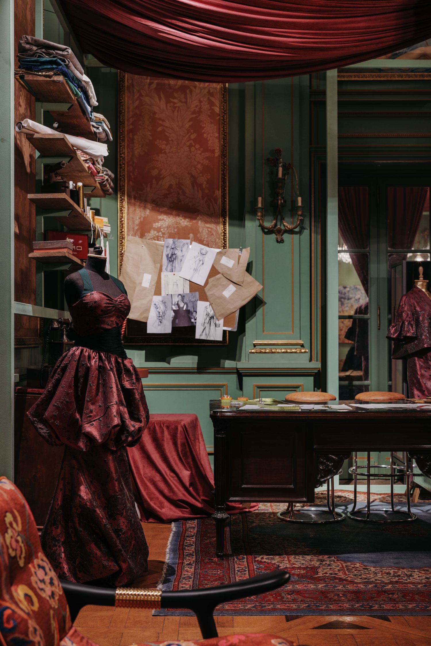 Elegant burgundy gown on mannequin in a vintage fashion studio with green walls and design sketches pinned nearby.