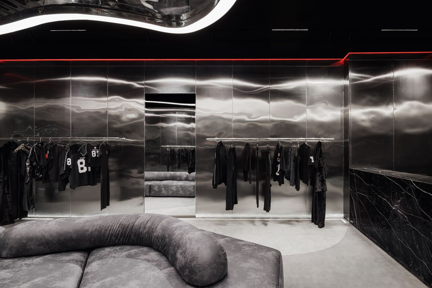 Dark luxury fashion boutique with metallic wave walls, gray velvet sofa, and black clothing displays under curved LED lighting.