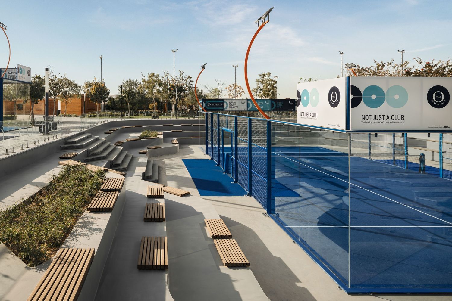 Modern outdoor sports complex featuring blue padel courts, wooden benches, and a skate area under clear sky.