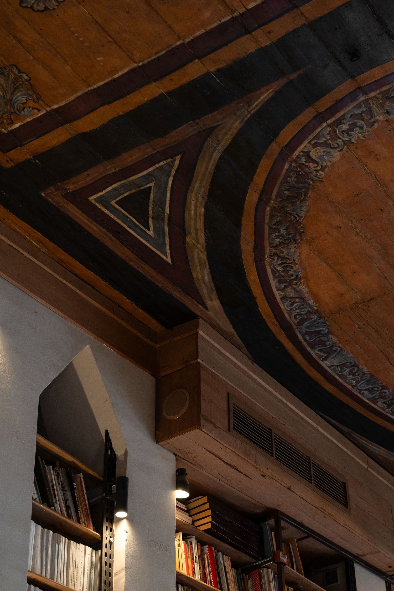 Close-up of an ornate painted wooden ceiling with geometric triangular motifs above a warmly lit bookshelf.