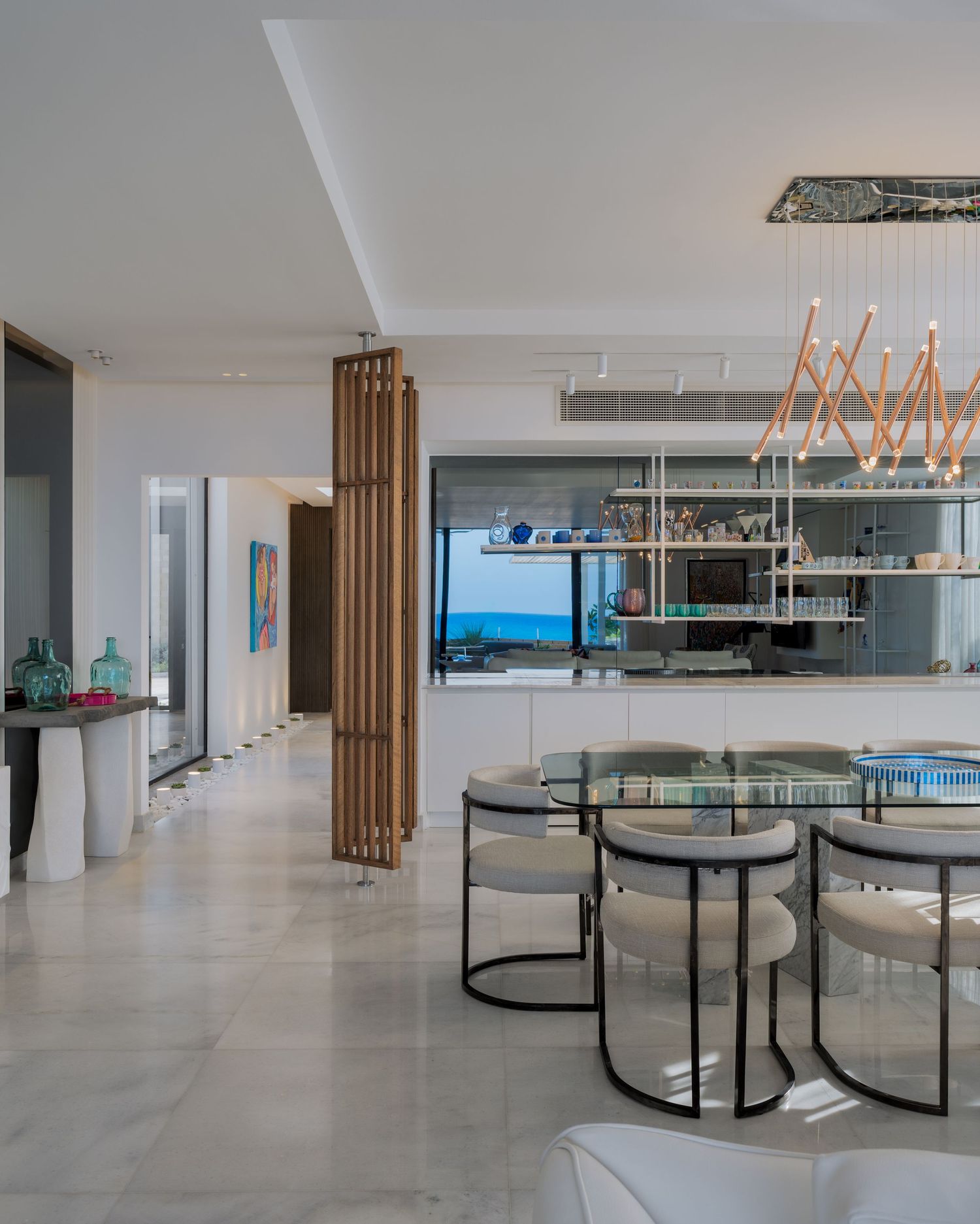 Modern open-plan dining area with glass table, white shelving, and ocean view beyond wooden partition.